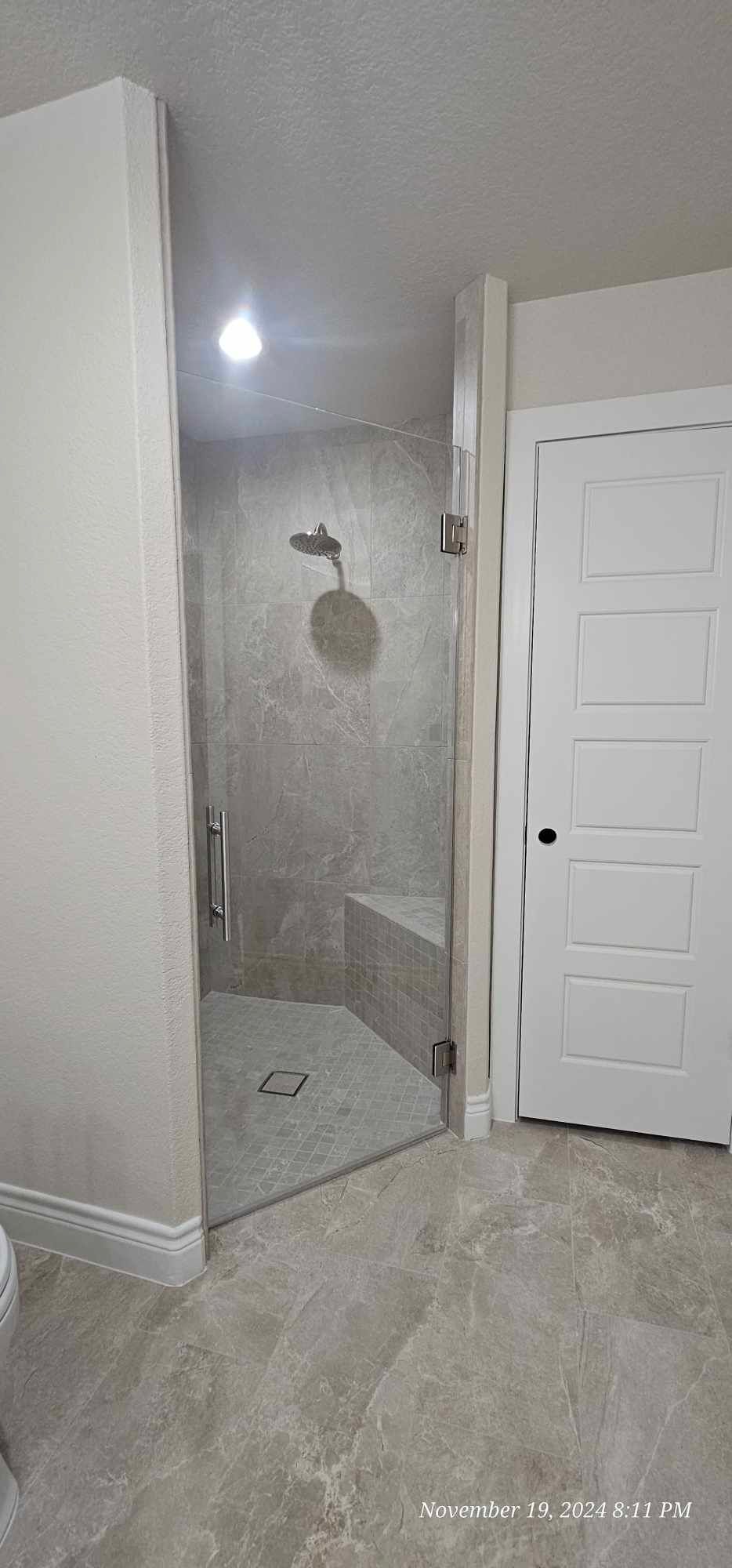 A modern bathroom with a glass shower enclosure and tiled flooring. A white door is on the right.