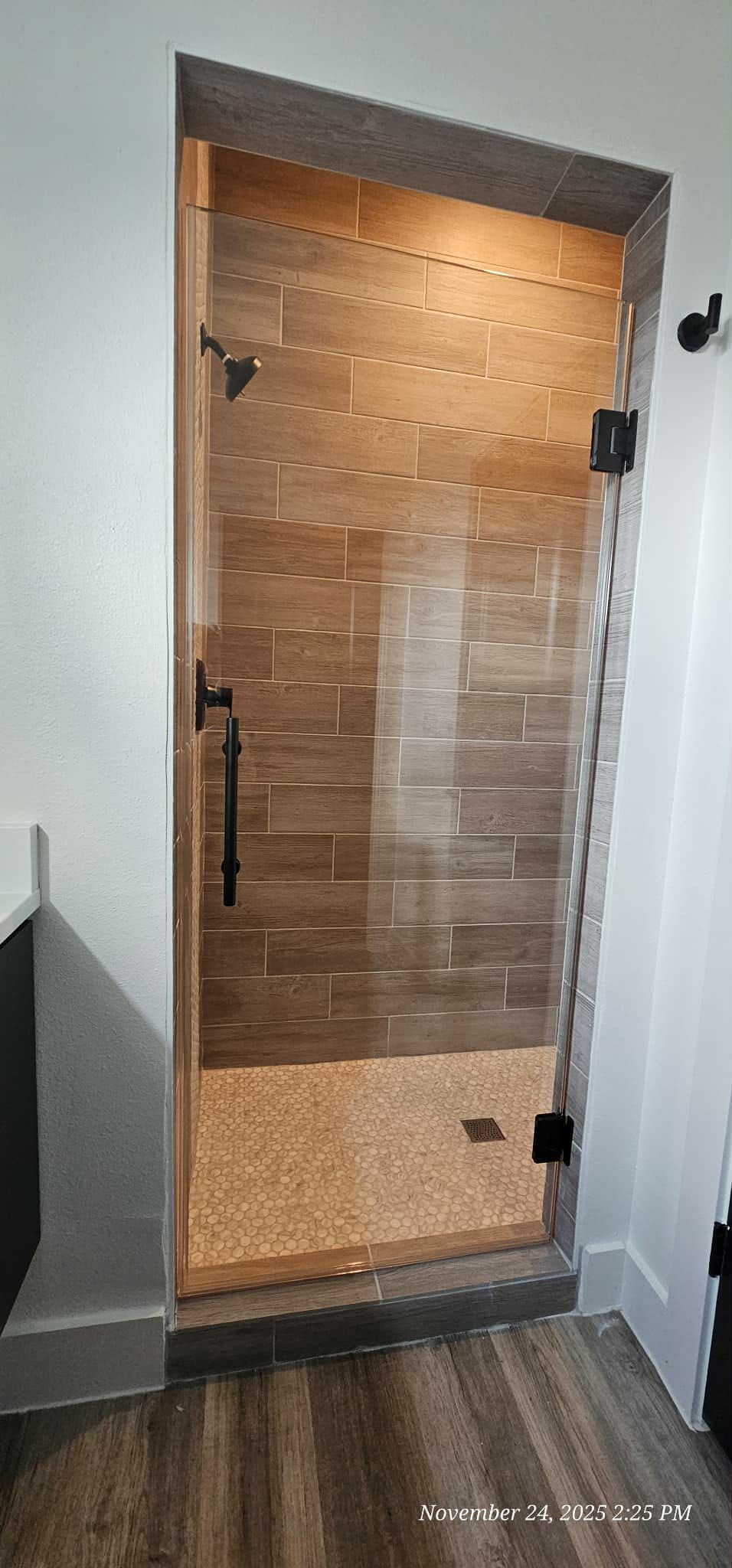 A modern shower with a glass door, dark fixtures, and wood-like tile.
