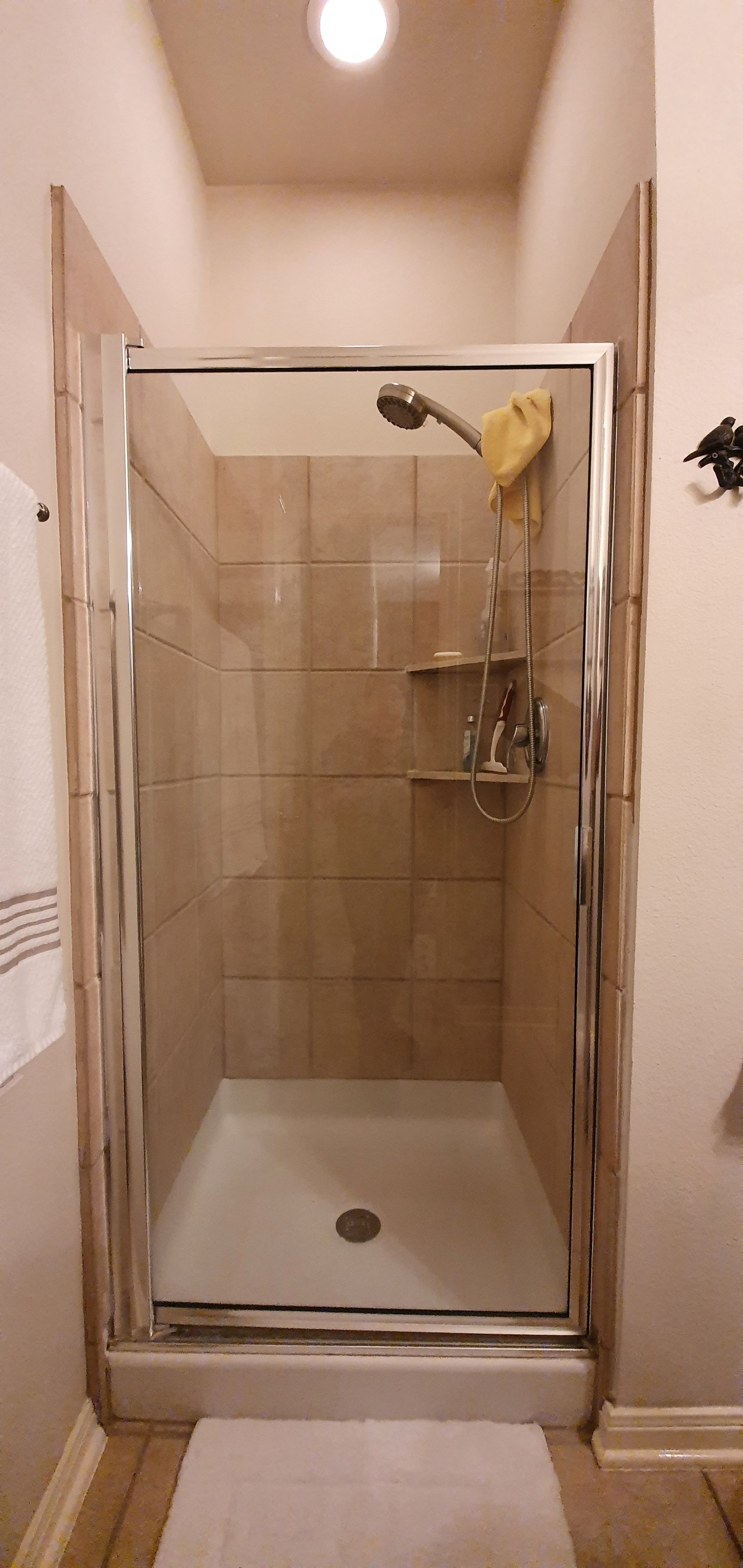 A shower with glass doors, brown tiled walls, and a white shower basin. A white bath mat is in front.