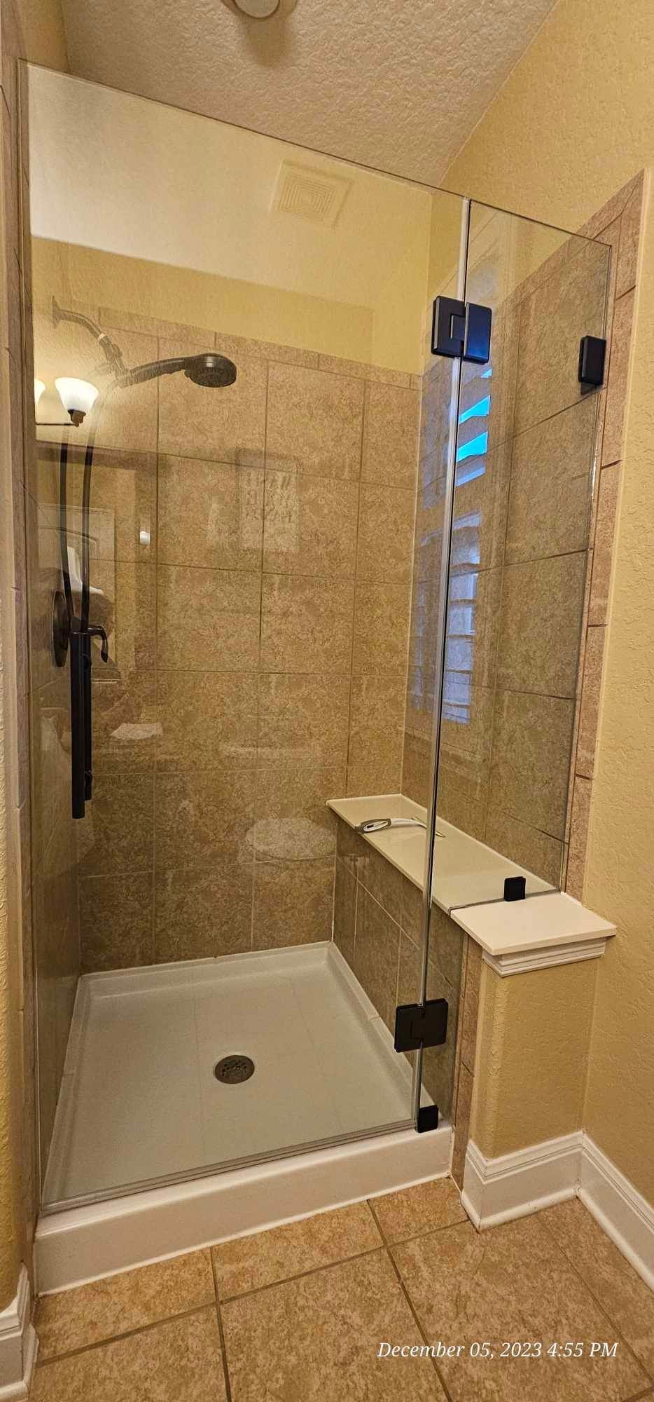 A tiled shower with a glass door, showerhead, and a bench. The shower stall is beige.