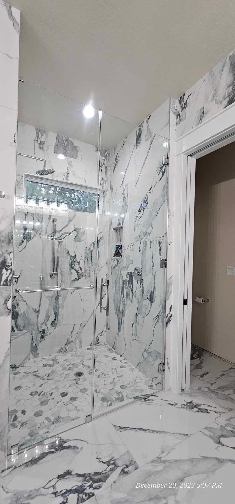 A marble-tiled shower with glass doors, silver fixtures, and a recessed window in a bathroom.