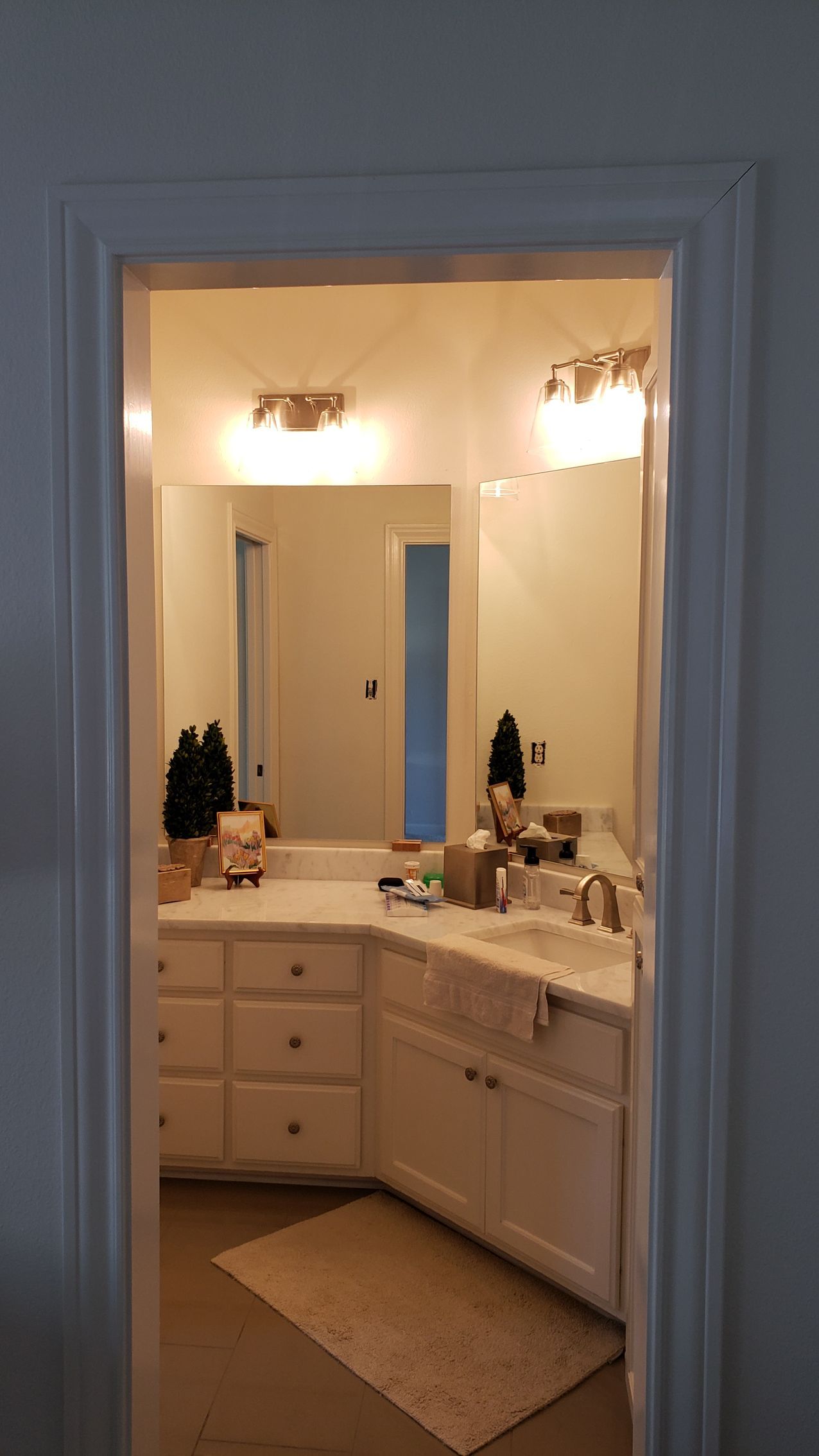 Bathroom viewed through a doorway, with white cabinets, lights, mirrors, and beige rug.