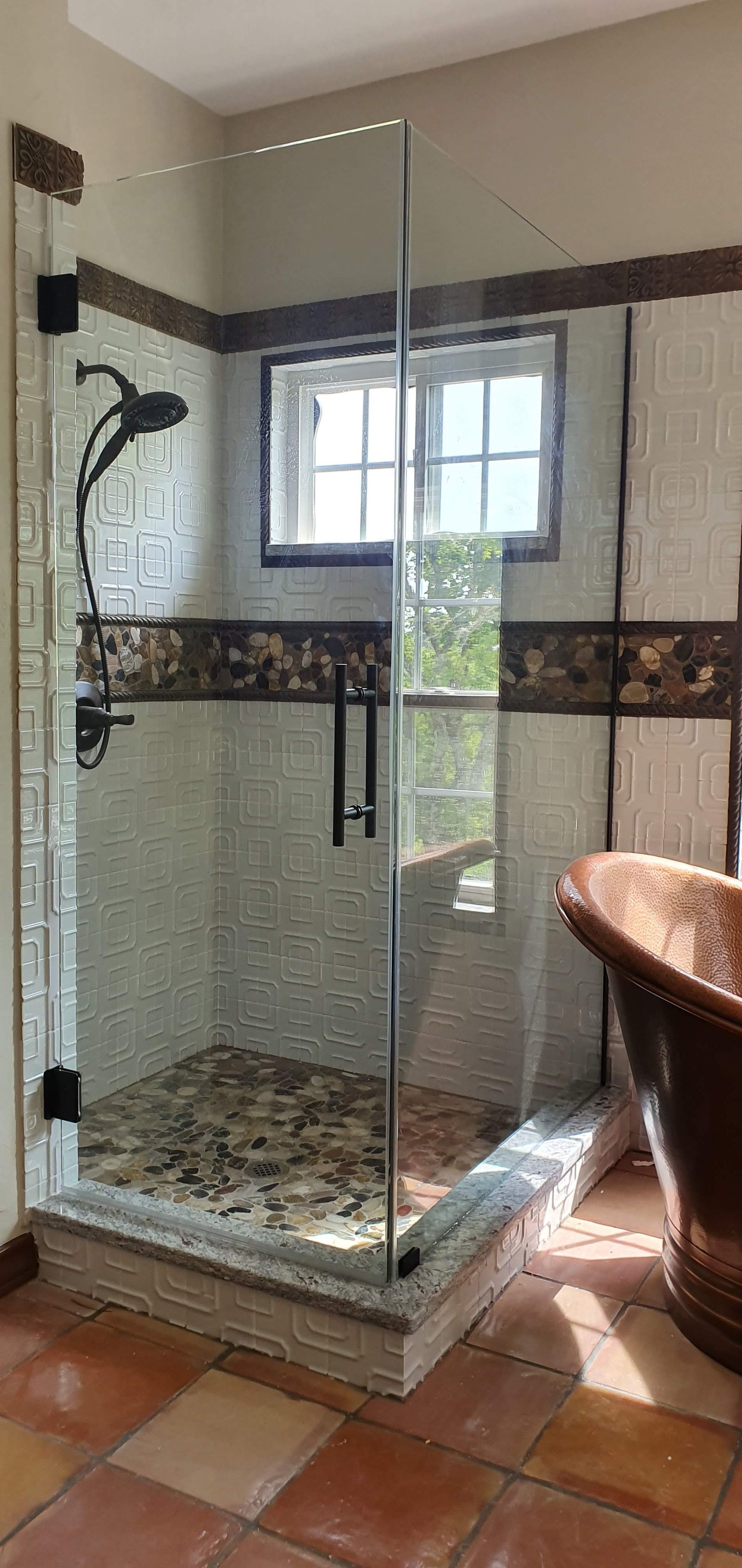 A corner shower with a stone floor and glass doors in a bathroom with a copper tub.