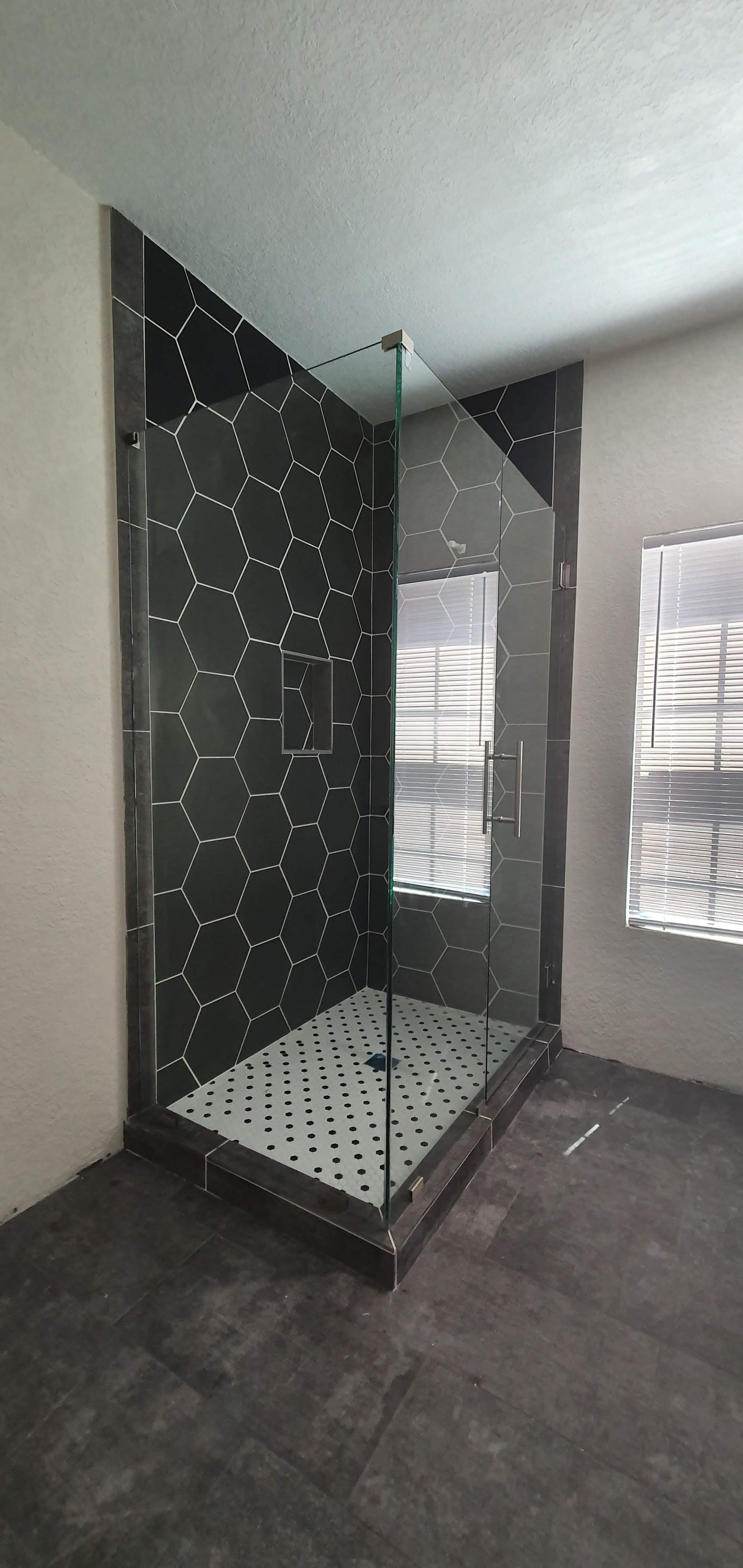 A newly constructed shower with dark hexagon-patterned tiles and glass doors, inside a room with a window.