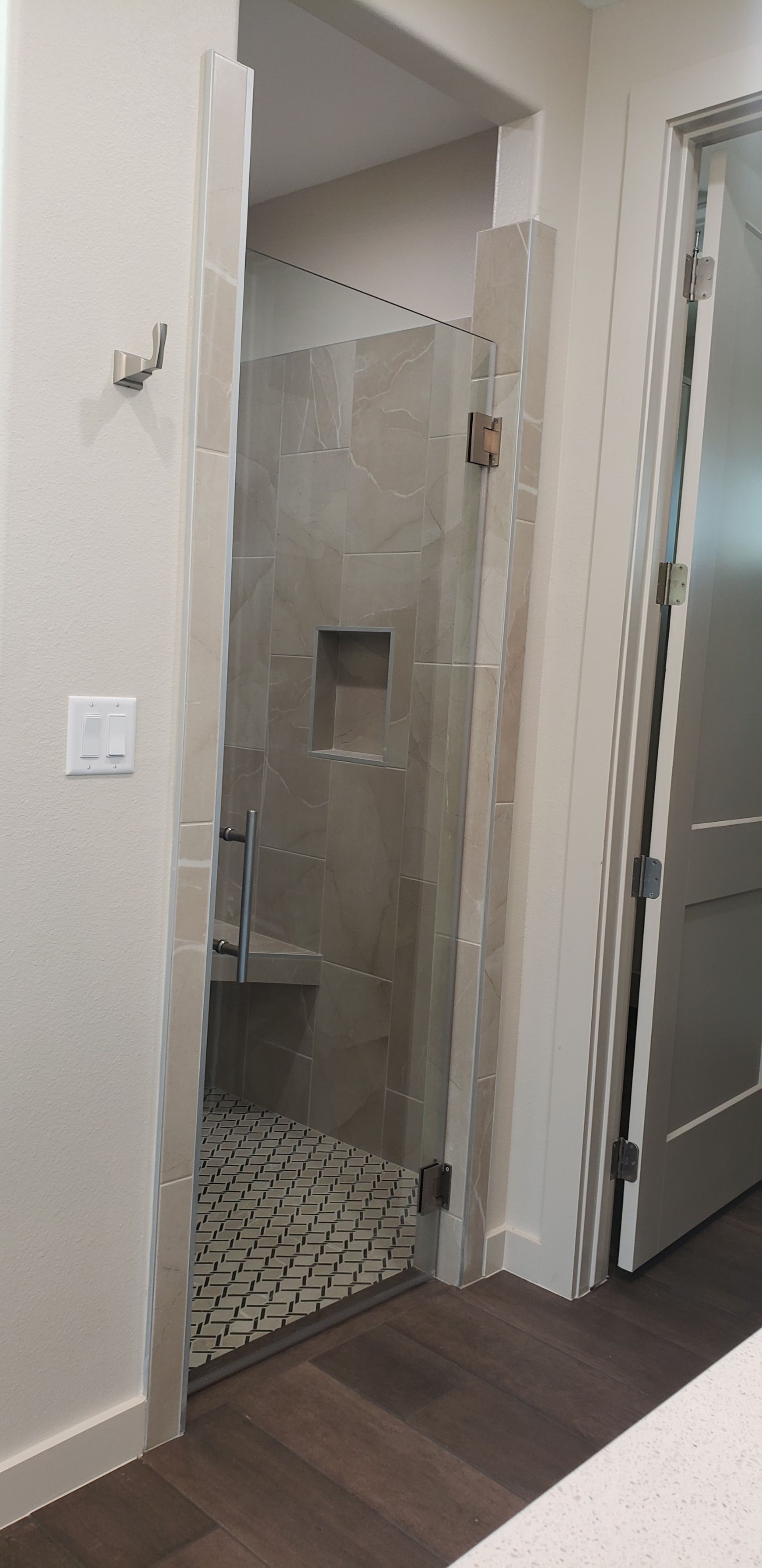 A bathroom with a shower. The shower has a glass door and gray tiled walls with a built-in shelf. Dark wood floor.