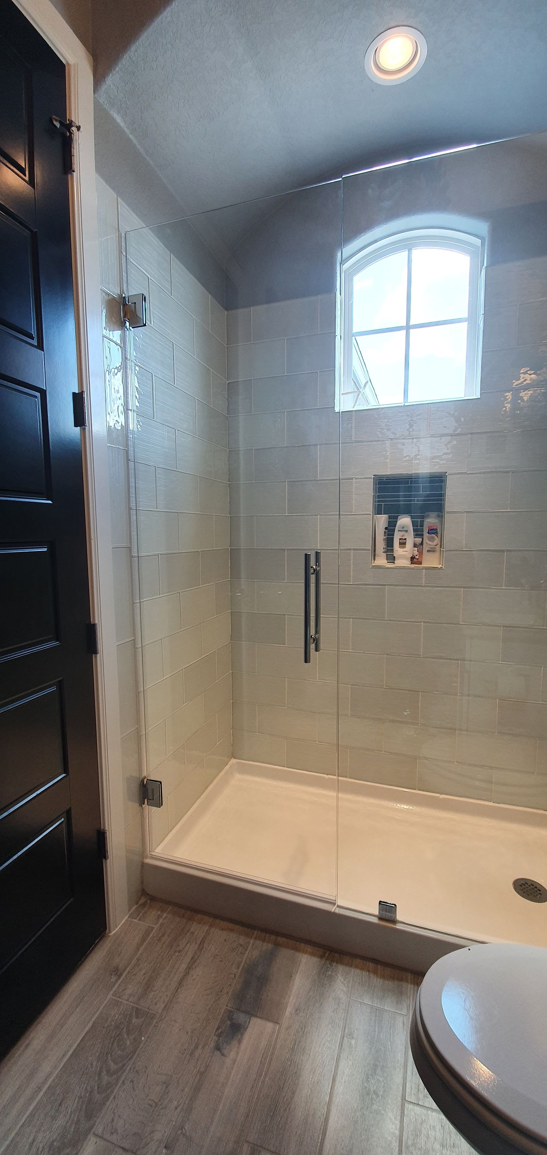 Shower with glass door, white tiled walls, and arched window. Dark wood door and wood-look floor.