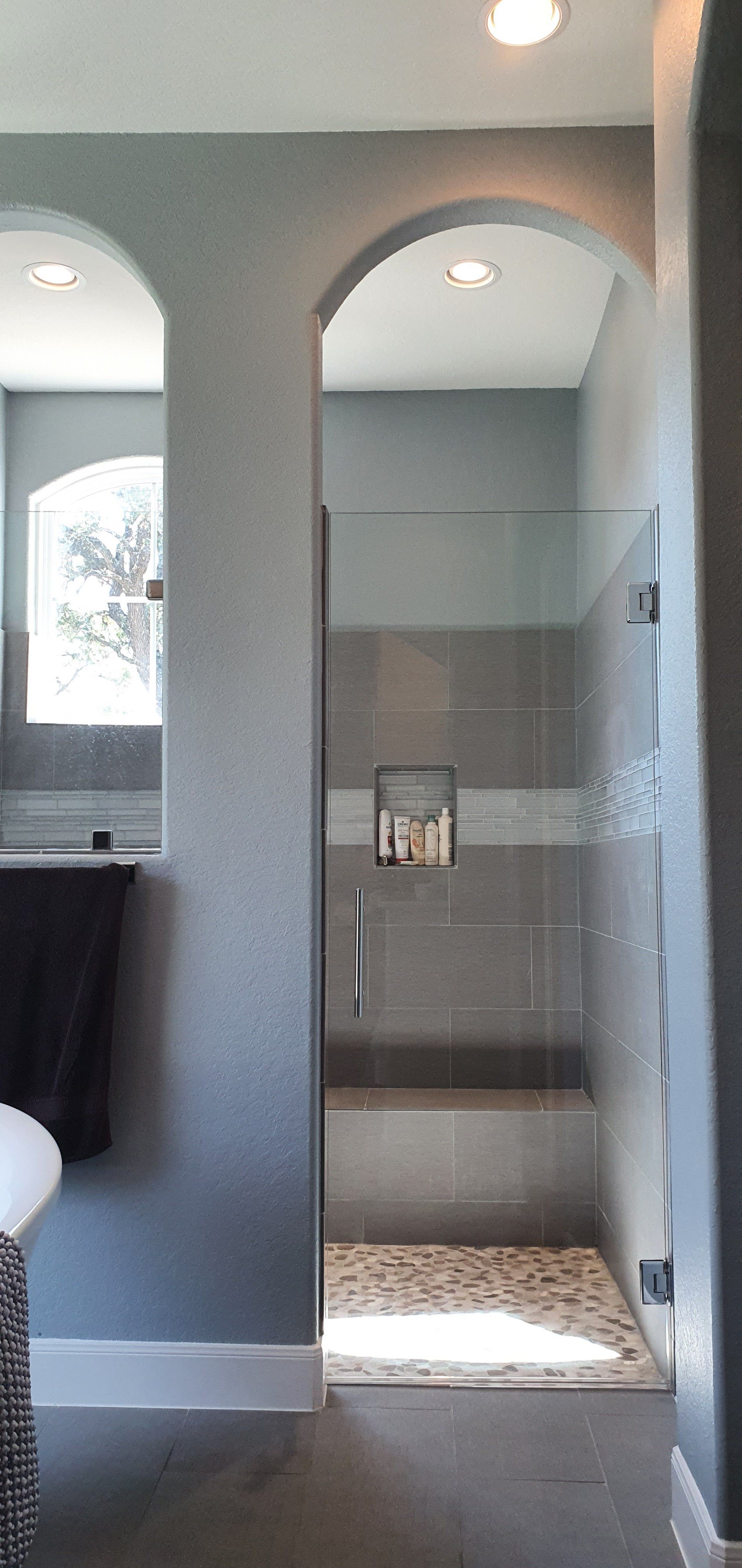 A modern bathroom with a glass shower and archways. Gray walls, light-colored stone floor, and a white trim.