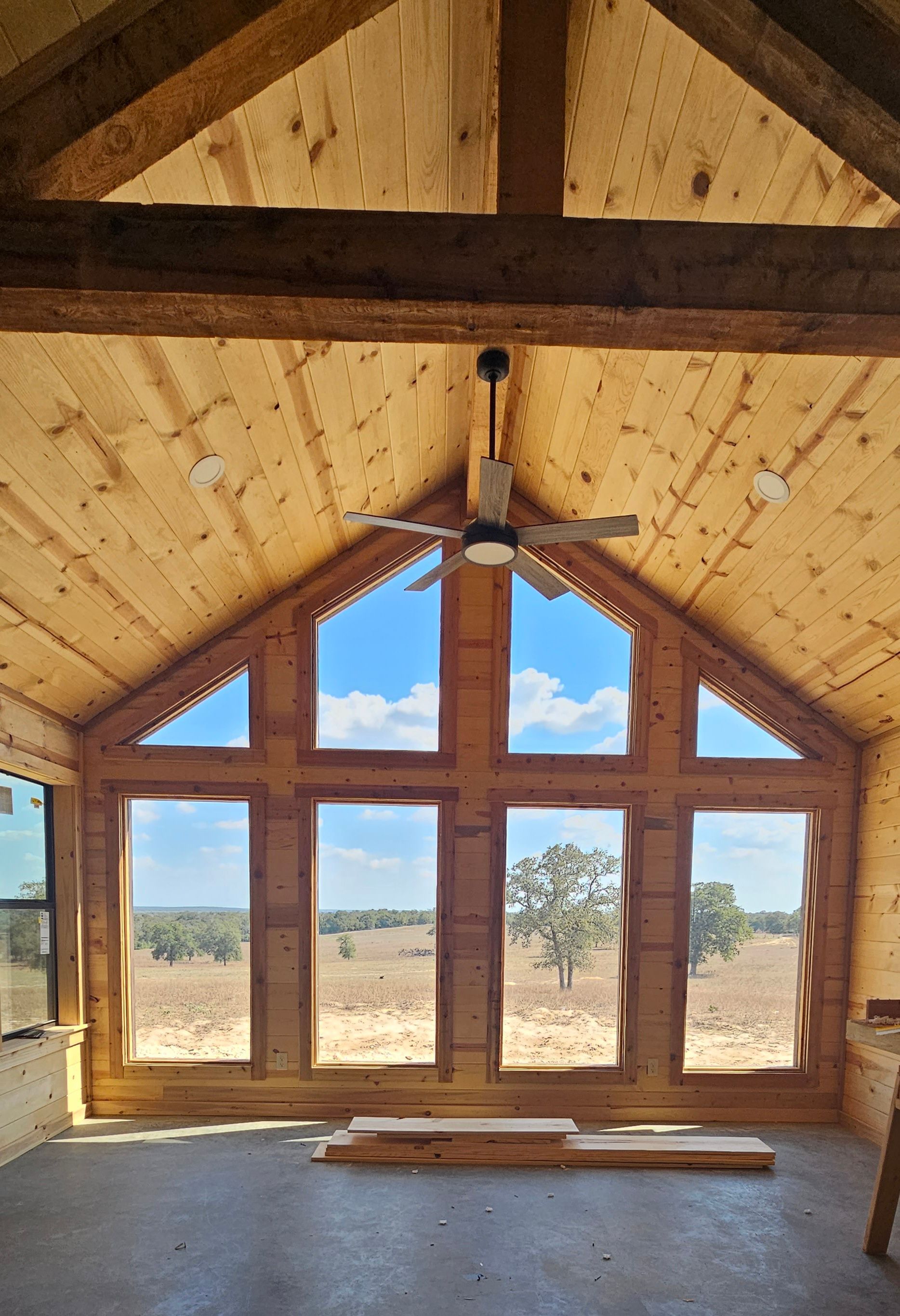 Interior of wooden structure with large windows overlooking a field, blue sky visible. Ceiling fan hangs from the roof.