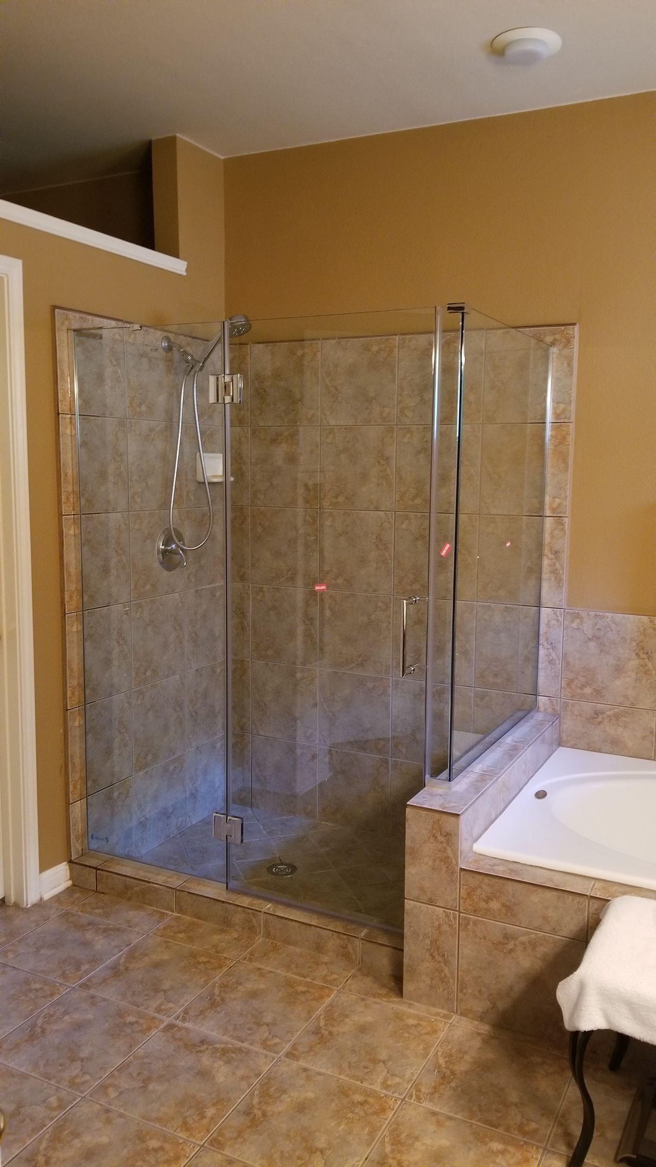 Glass shower with tan tile walls and floor, adjacent to a built-in tub.