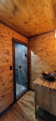 Rustic bathroom with wooden walls, ceiling, and floor. Shower door, vanity with a bowl sink.