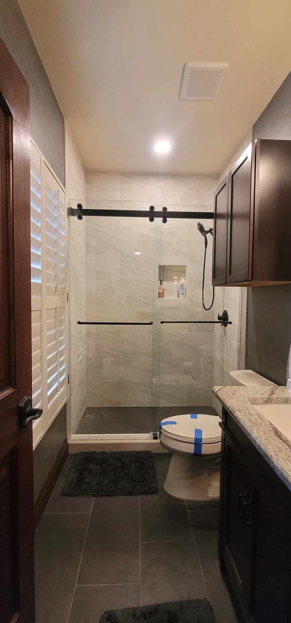 Bathroom with glass shower, toilet, dark cabinets, dark tile, and window with blinds.