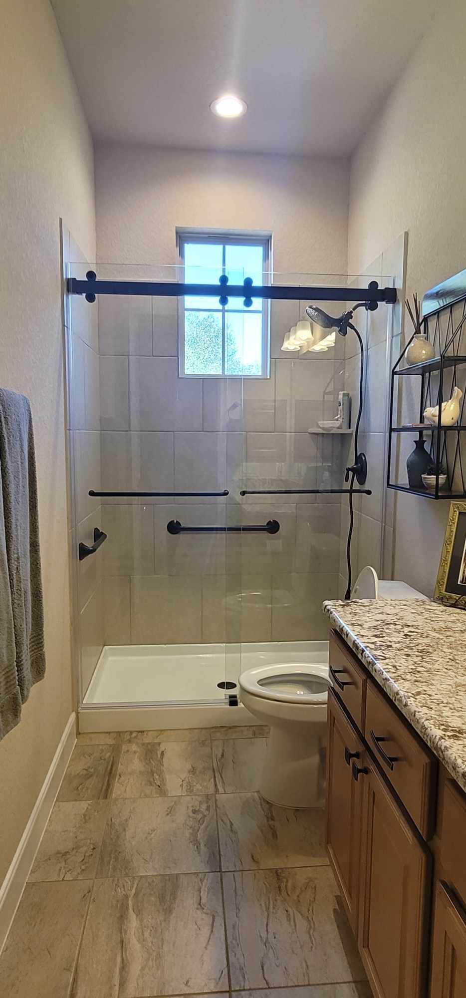 Bathroom with shower, toilet, vanity, and decorative shelving. Light beige walls, stone-like floor tiles, glass shower door.