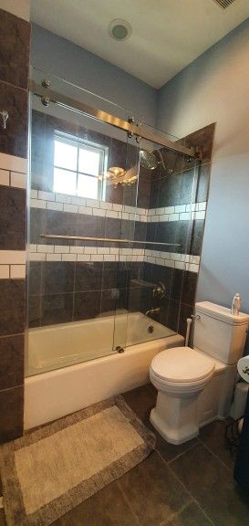 Bathroom with a white bathtub, toilet, and brown tiled walls. A shower door and a rug are present.
