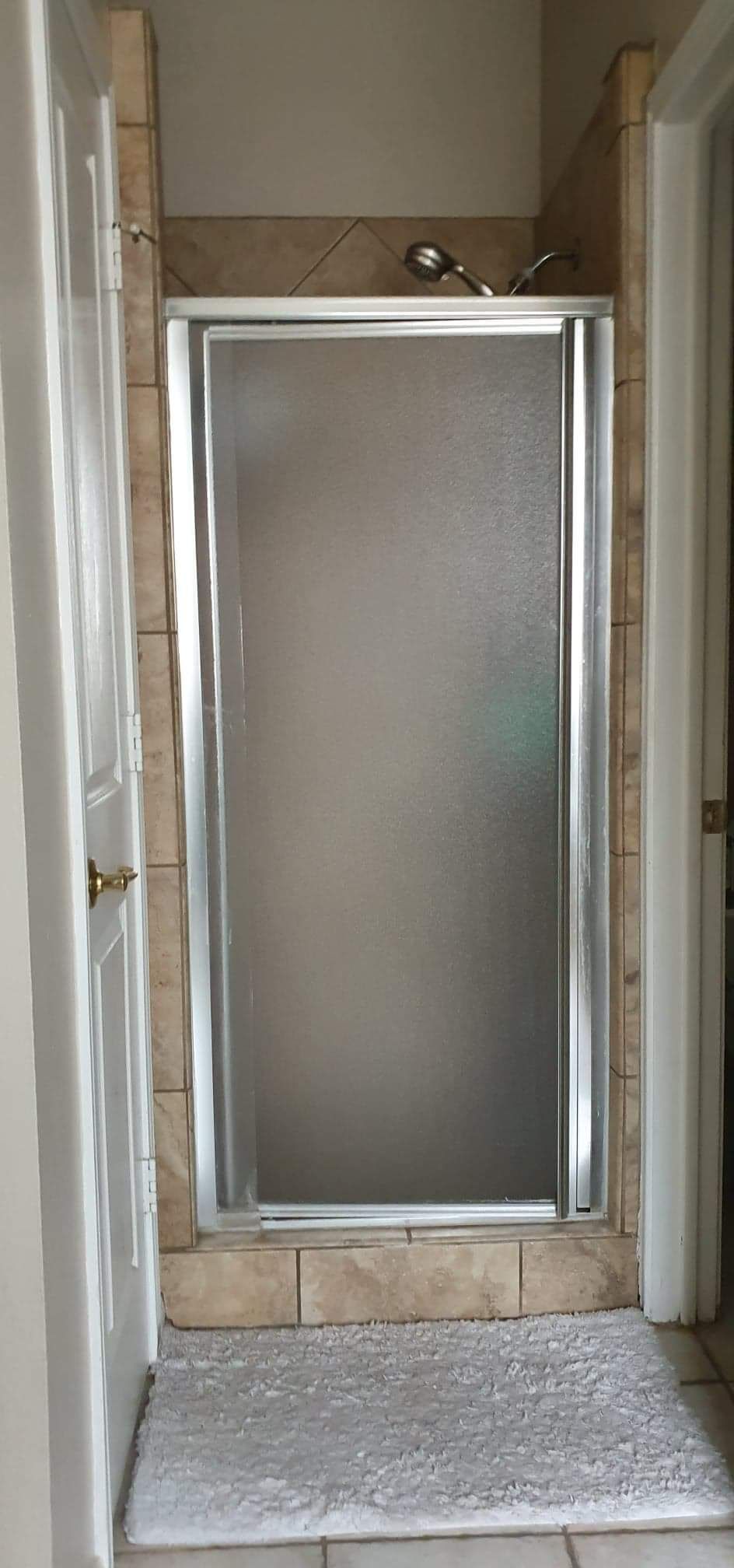 Shower stall with frosted glass door, set between tiled walls and a door on either side. A bath mat is on the floor.