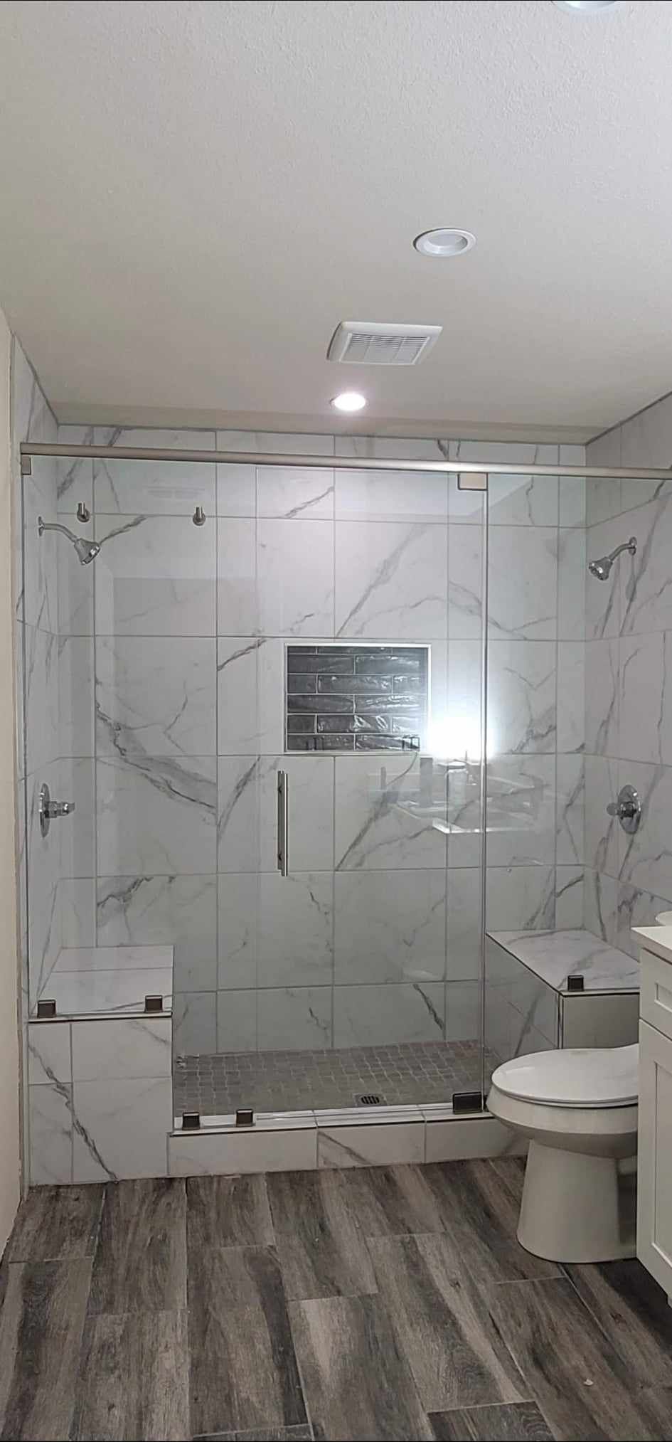 A modern bathroom with a glass shower and marble tile walls. Gray wood-look floor, toilet, and white vanity.