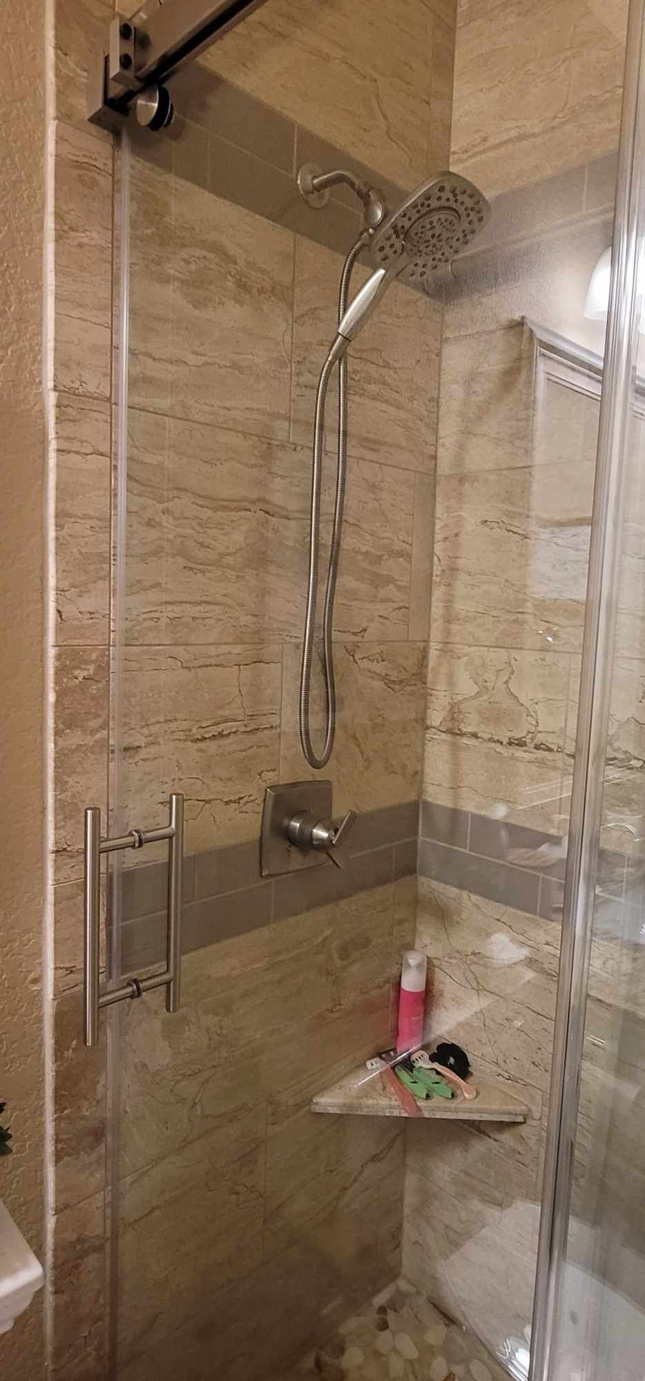 Shower with glass door, silver fixtures, and beige tiled walls. A shelf holds toiletries.