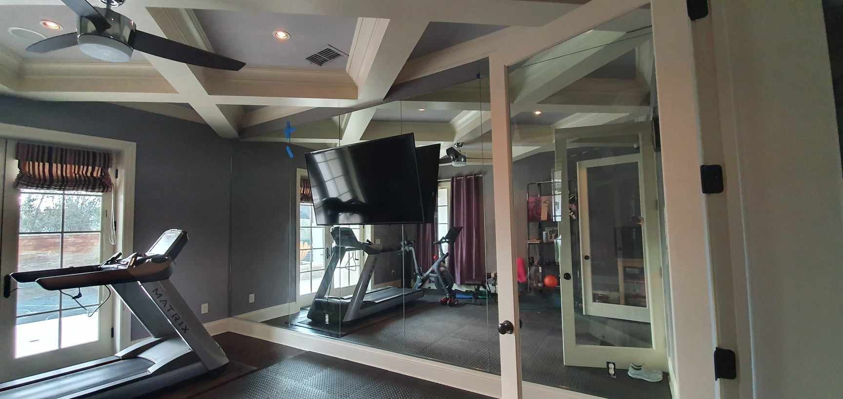 A home gym with treadmills, a TV, windows, and mirrored walls. The ceiling has beams and a fan.