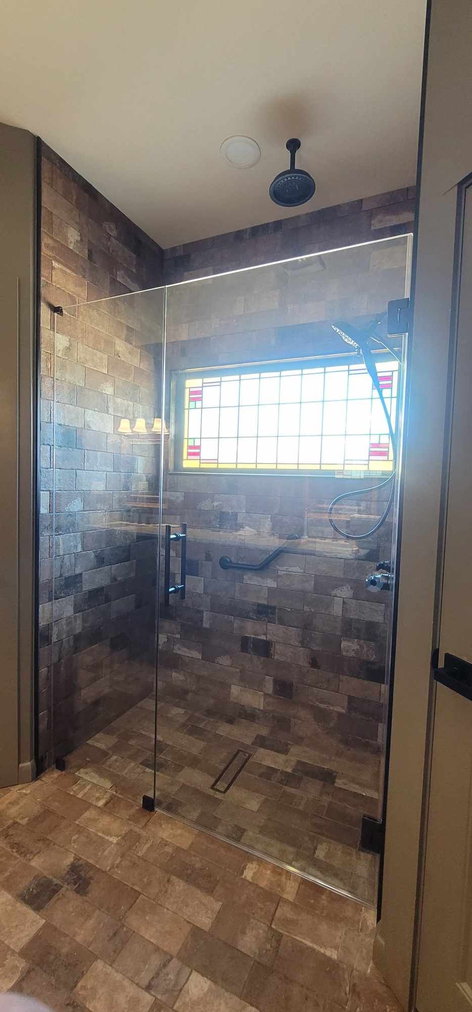A glass-enclosed shower with brown tile walls and floor, and a stained glass window.