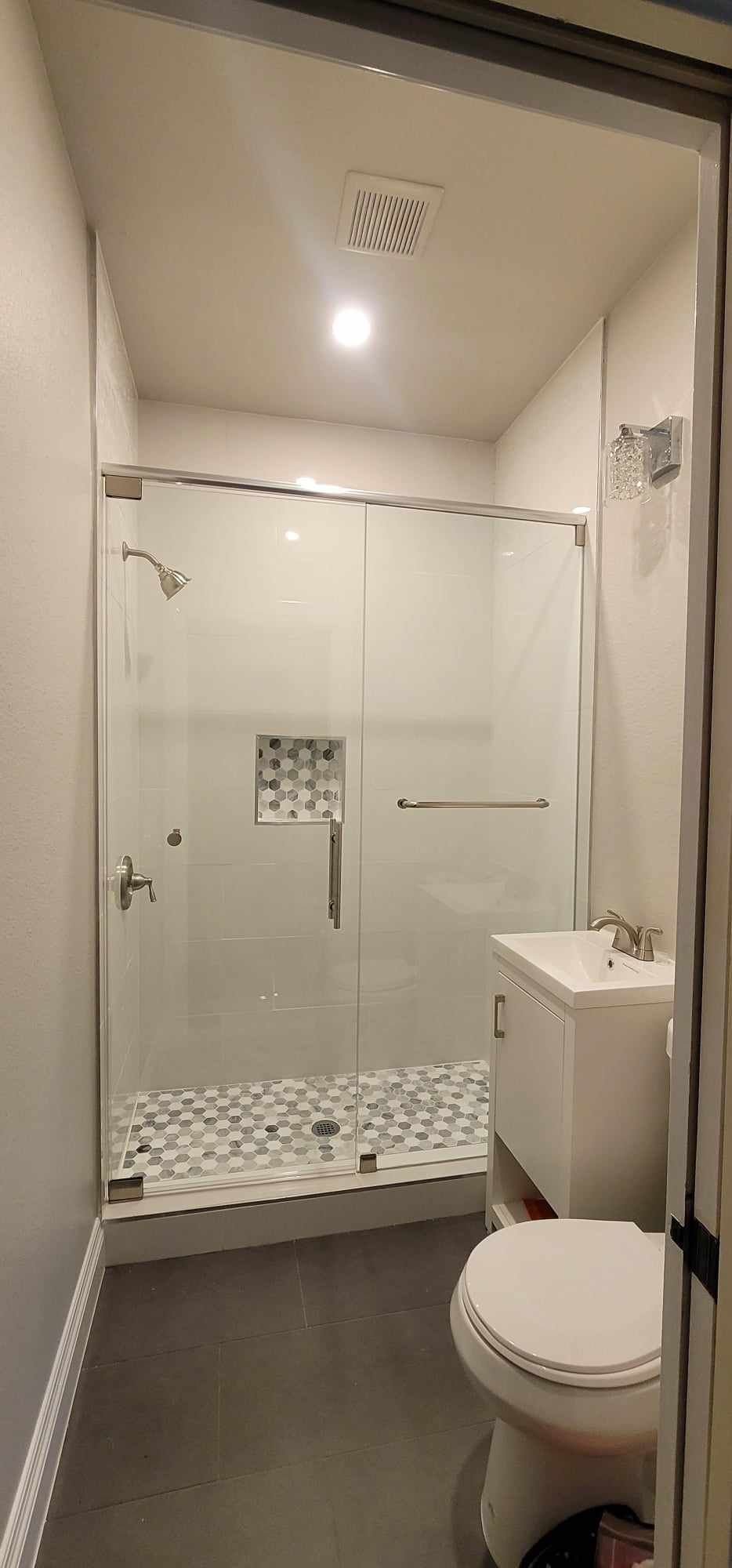 Bathroom with shower, vanity, and toilet; gray floor, white walls and fixtures, glass shower door.
