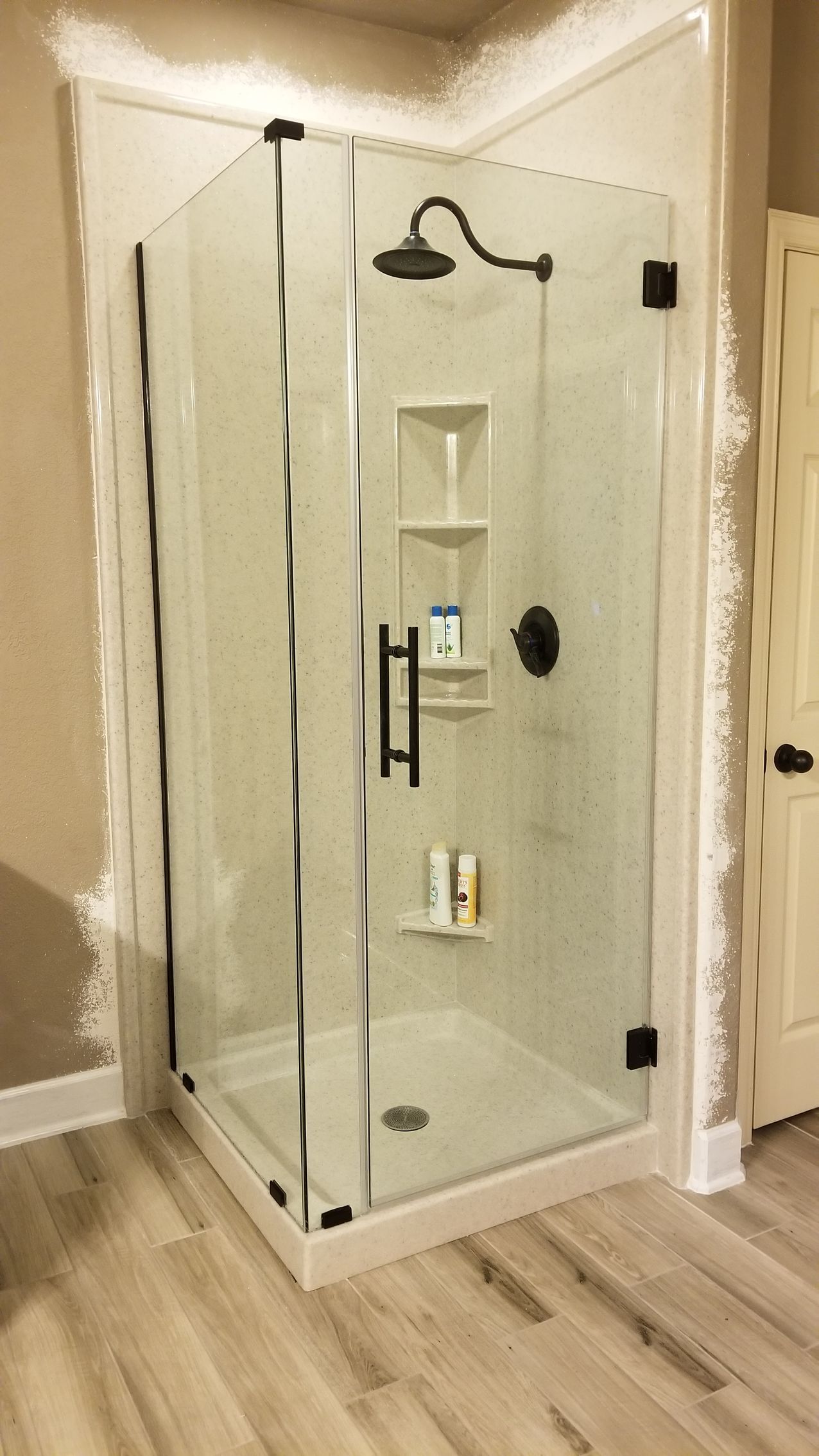 Corner glass shower with black fixtures and built-in shelving, on light wood-look floor.