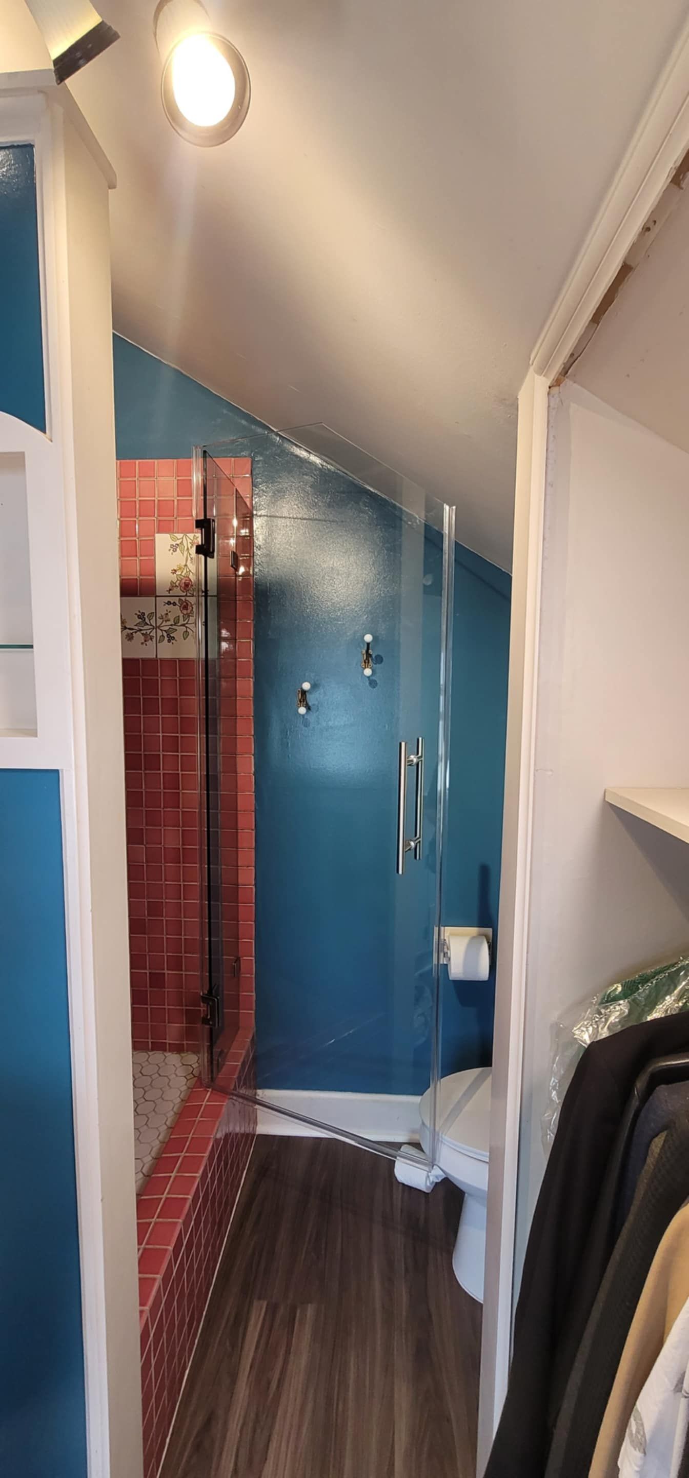 Narrow bathroom with blue walls, red tiled shower, glass door, and toilet. Wardrobe with clothes on the right.