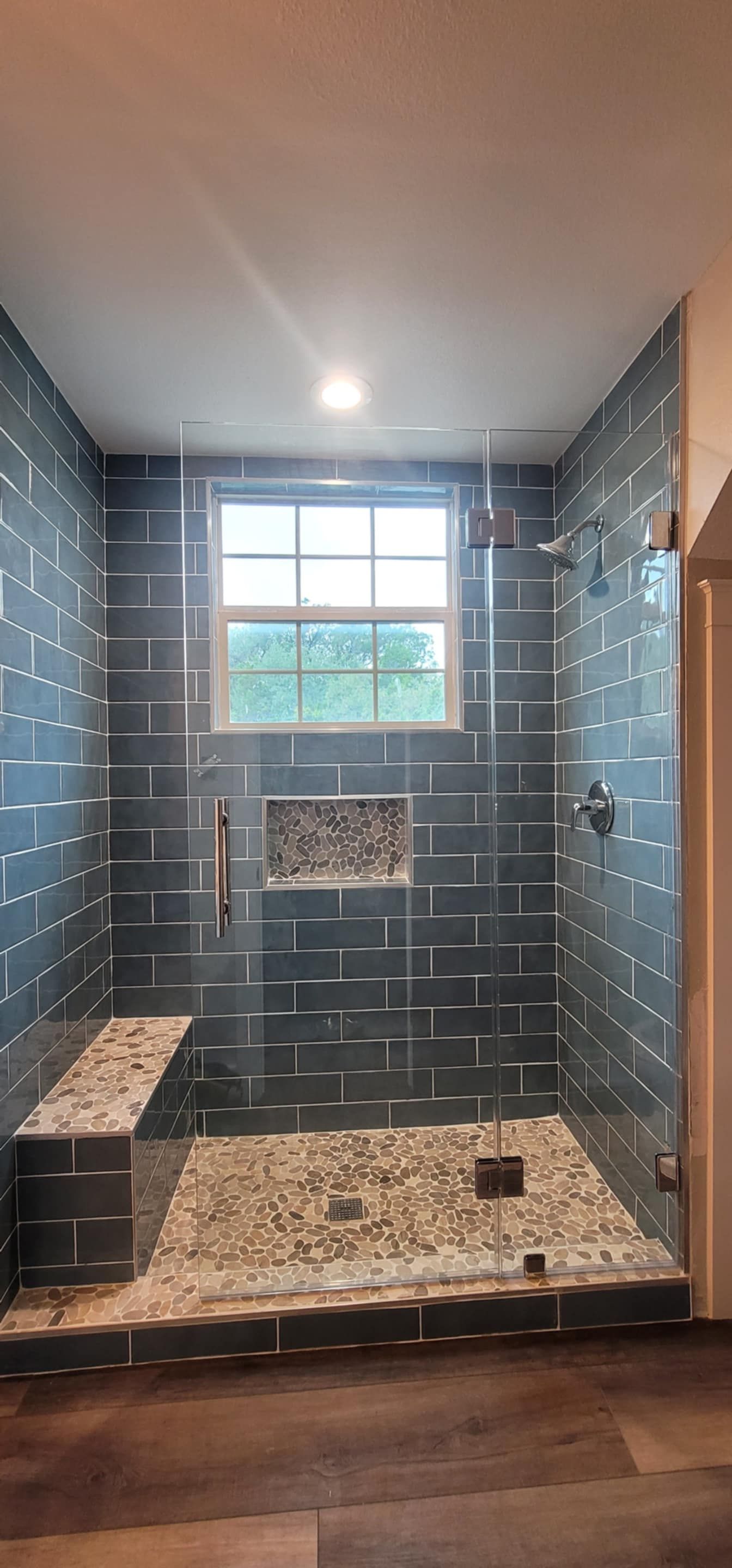 Shower with dark gray tiled walls, pebble floor, built-in bench, and a window.