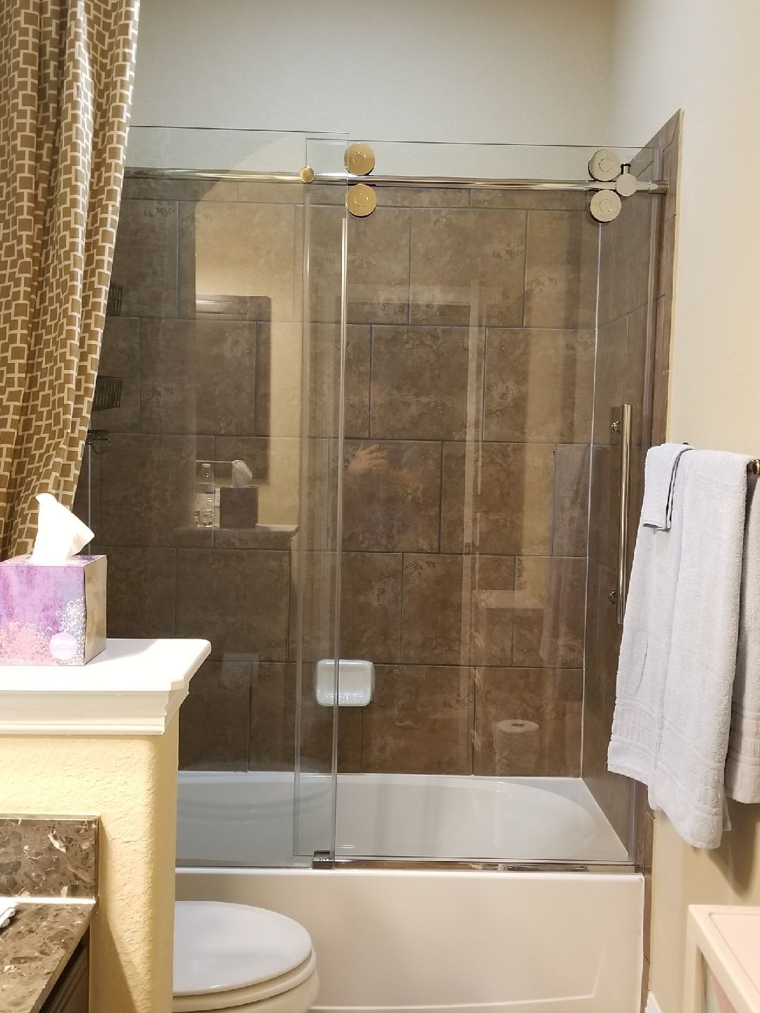 Bathroom with glass shower door, white tub, brown tile, beige walls, and light-colored towels.