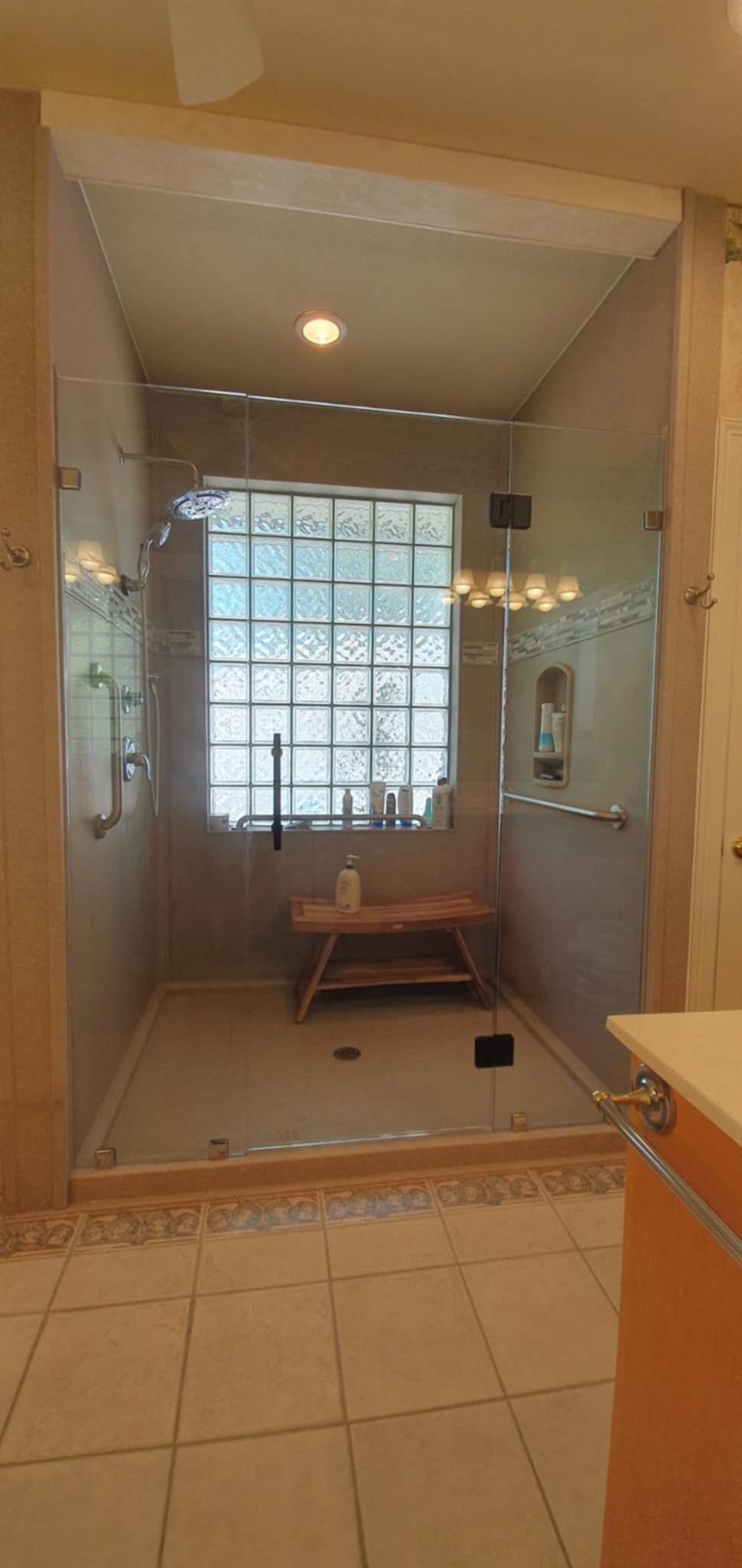 A glass shower with a small bench inside and a window behind it. Beige walls and floor.