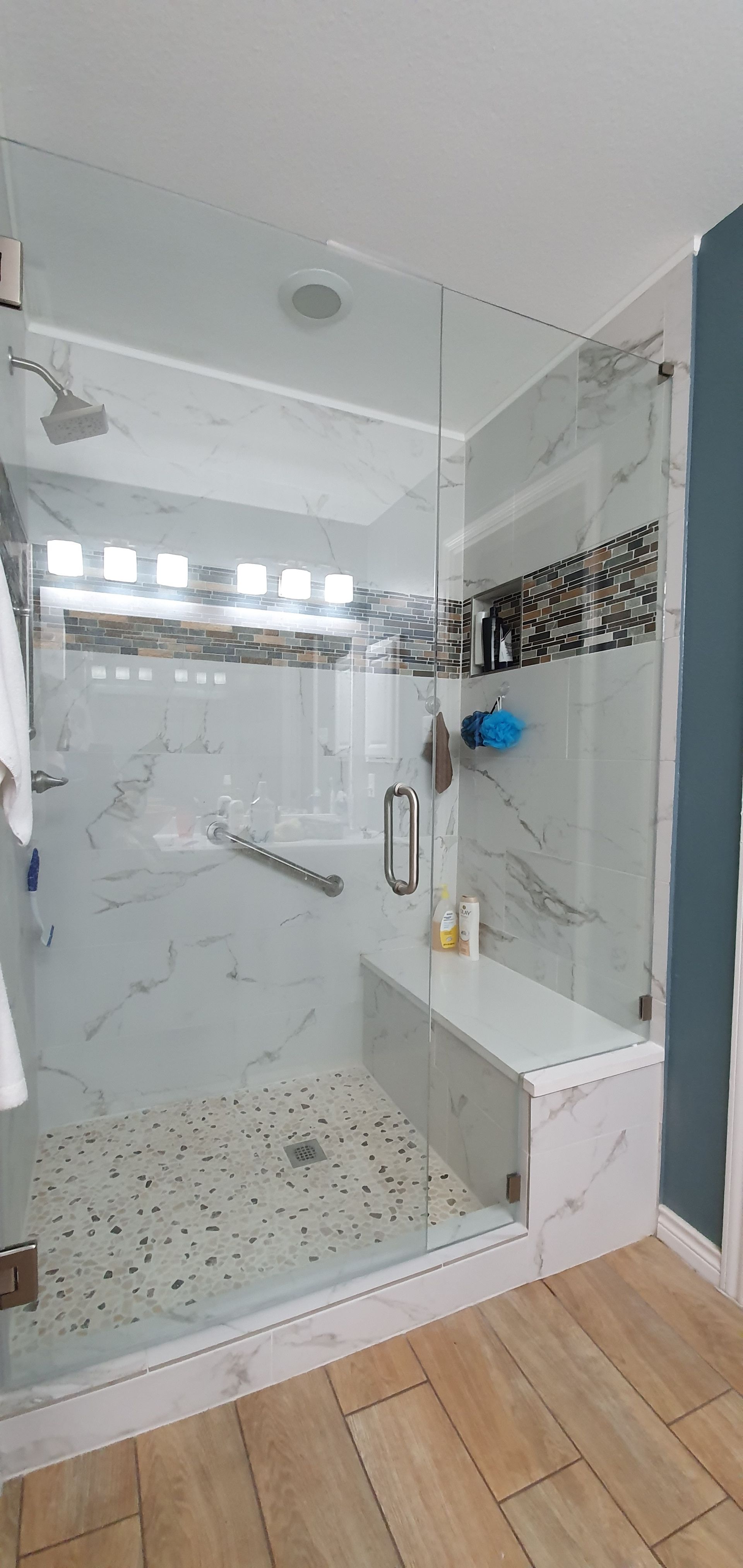 A modern shower with marble walls, a glass door, and a built-in bench. Wooden floor.