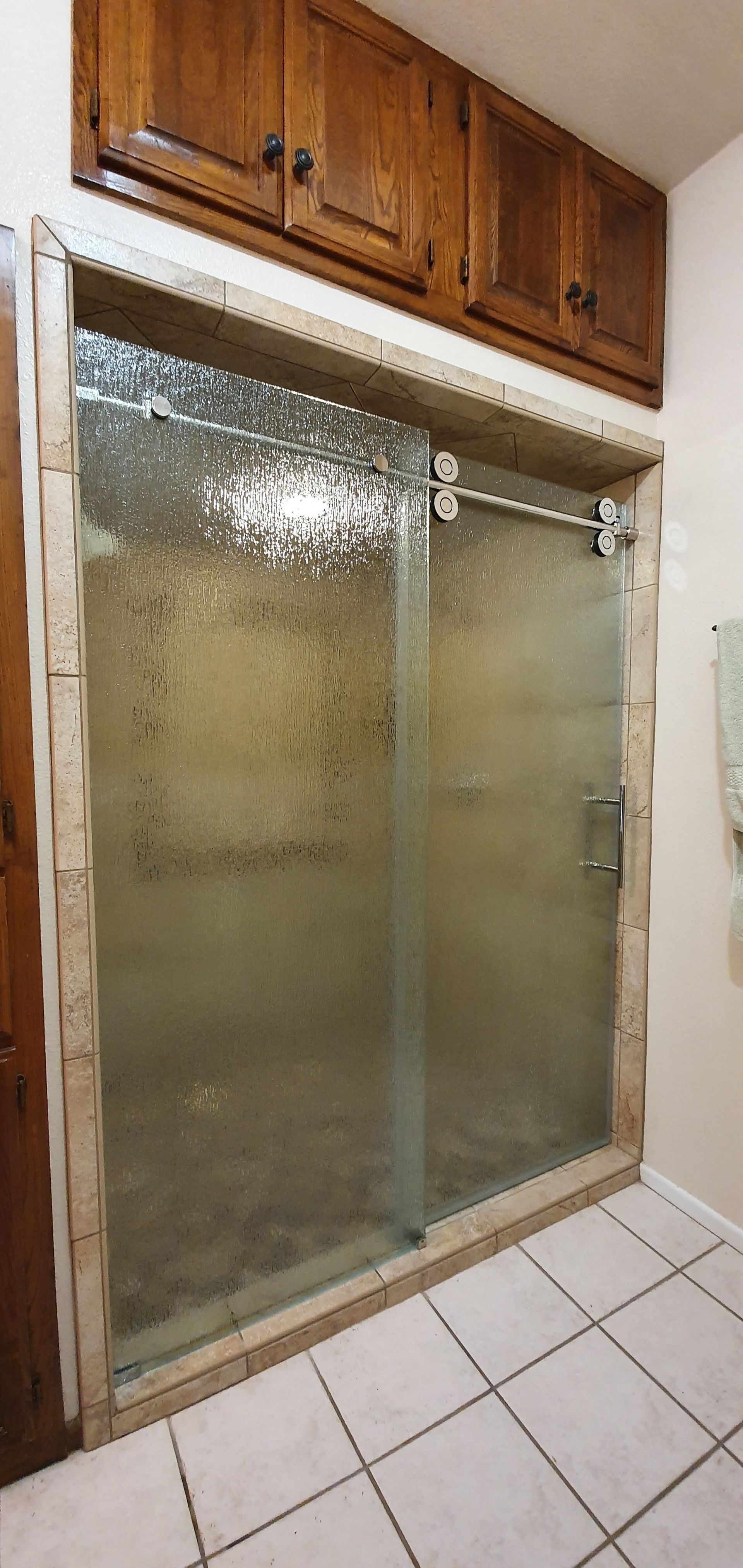 A bathroom shower with a glass door, framed with tan stone-like tiles, beneath wooden cabinets. White tile floor.