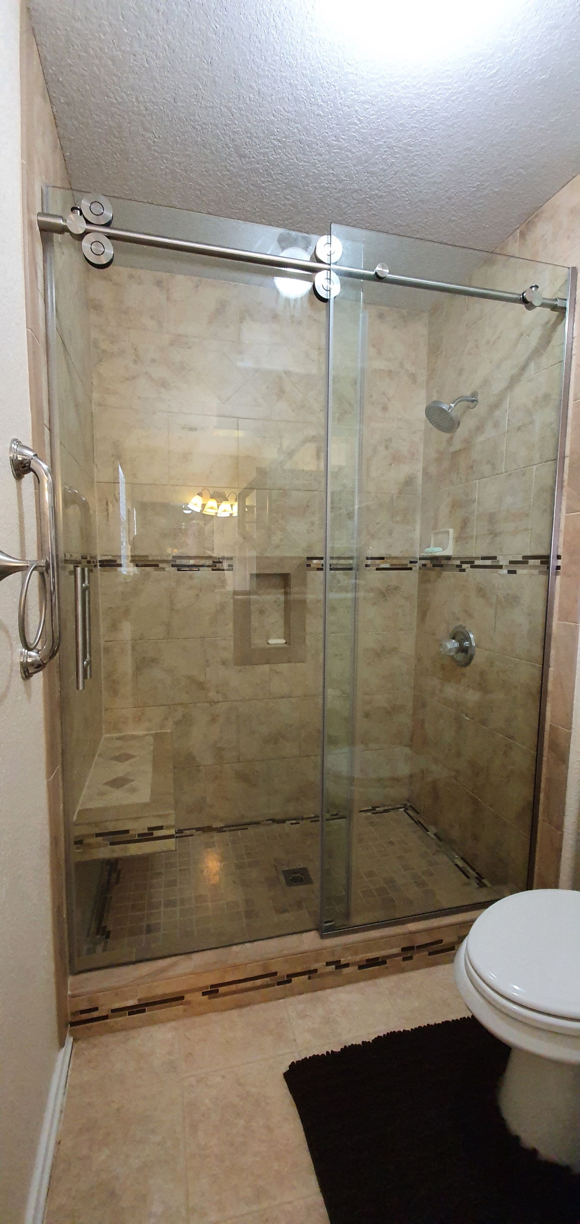 Bathroom with a glass shower enclosure and a toilet. Beige tile walls and floor. Black bath mat.