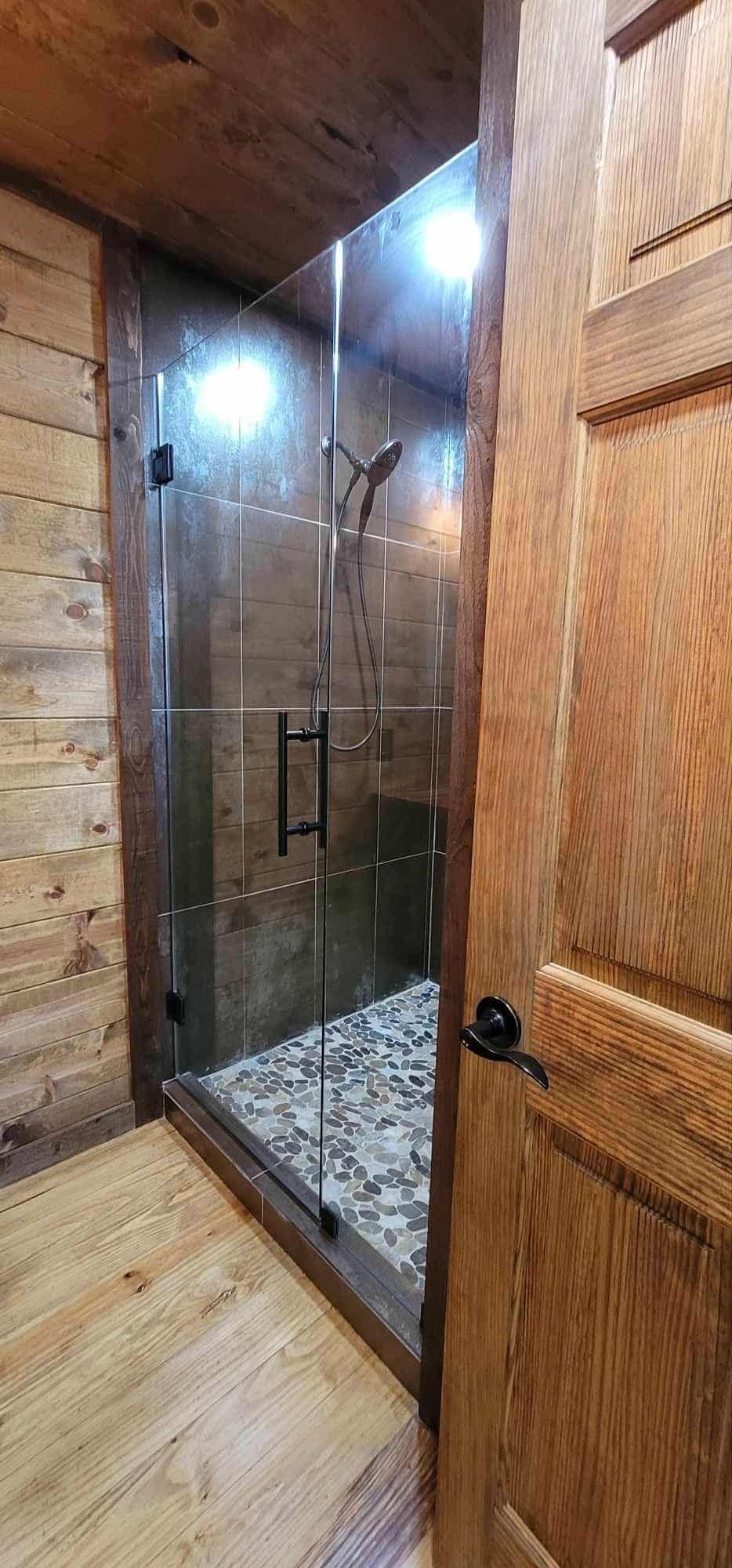A shower with a glass door, framed by wood, is visible from an open wooden door. The shower has stone flooring.