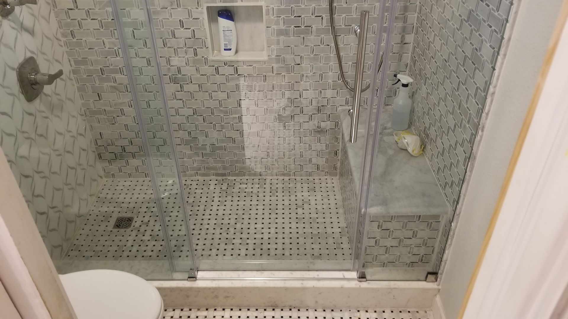 A shower with glass doors, patterned tile, and cleaning products.