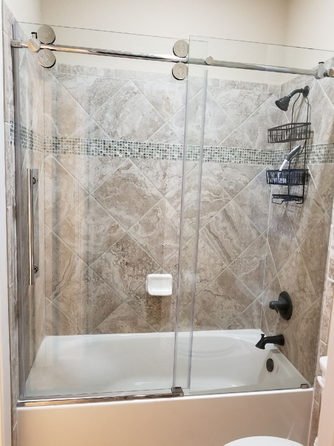 Shower with sliding glass doors, beige tiled walls, white bathtub, black fixtures.