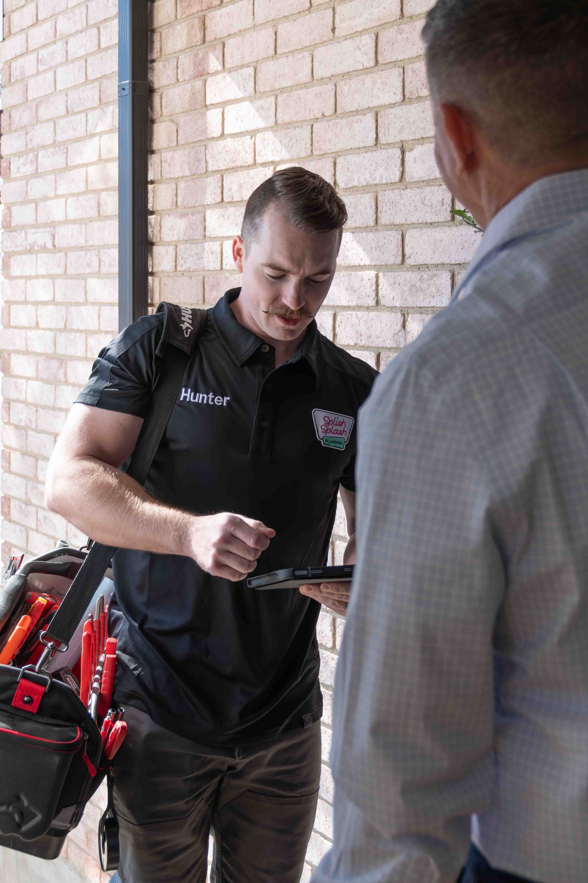 A man in a black shirt holding a tablet, speaking with another man near a brick wall, tool bag visible.