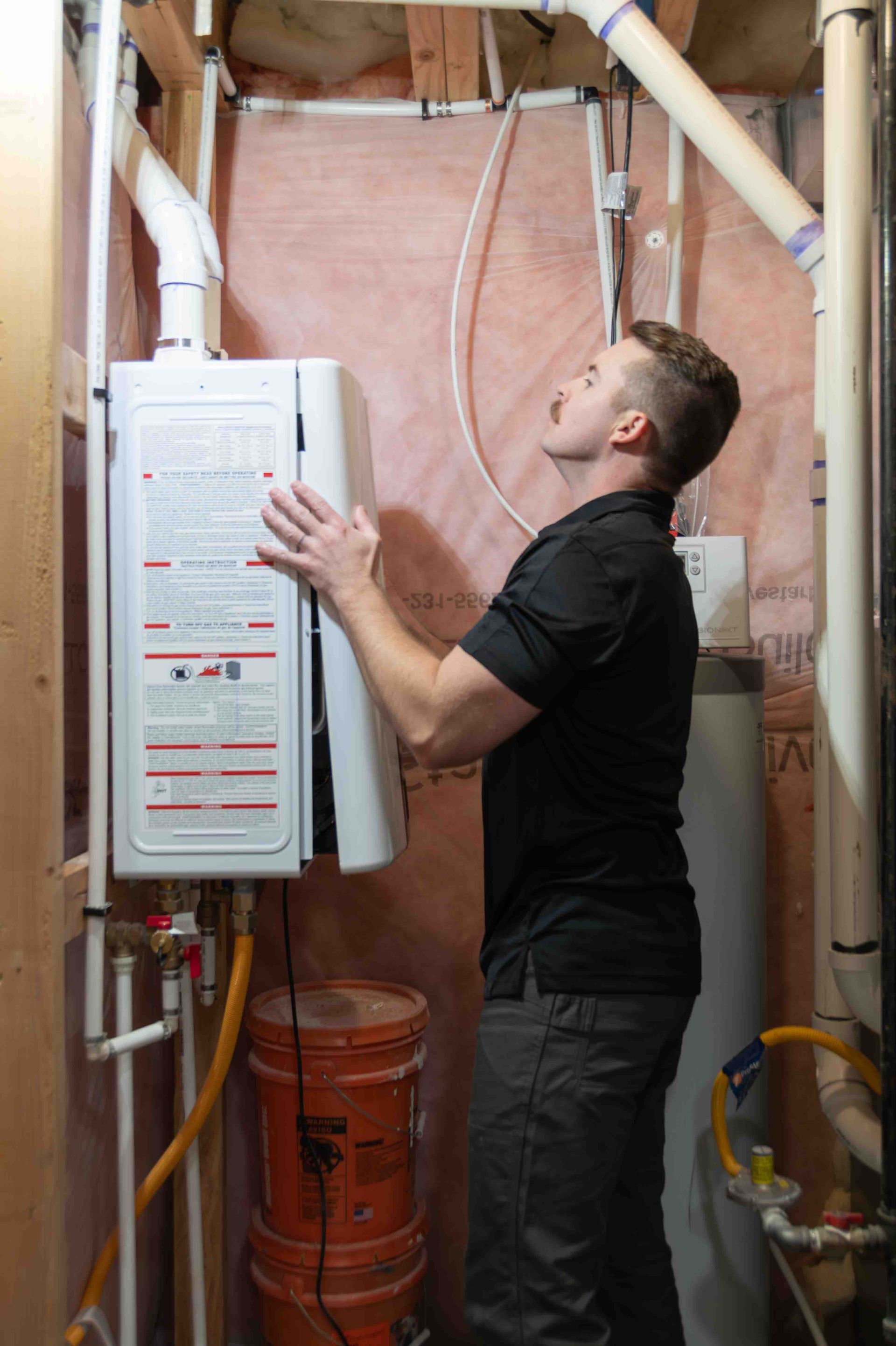 A person examines a white tankless water heater. The person wears a black shirt in a basement setting.