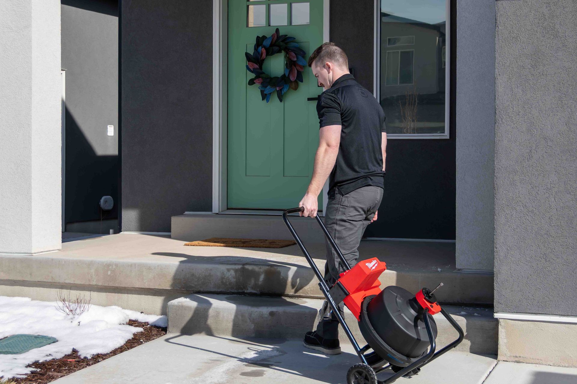 Man carrying a red and black plumbing snake machine up a porch towards a green front door.
