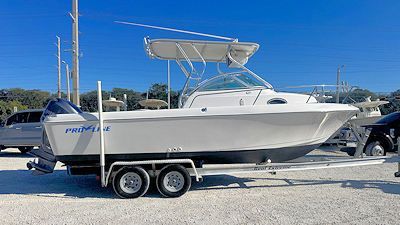 White boat on a trailer, with a blue motor and hardtop, parked on a gravel lot under a clear sky.
