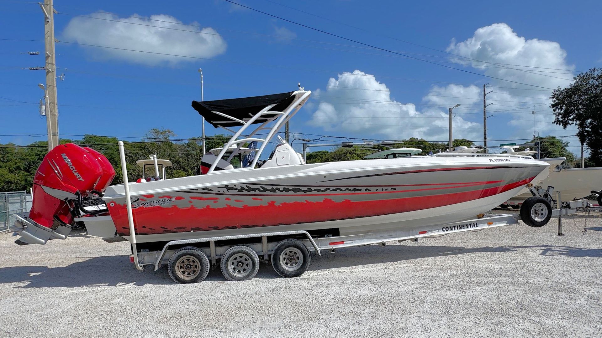 A white and red center console boat on a triple-axle trailer, parked on a gravel lot under a clear blue sky.