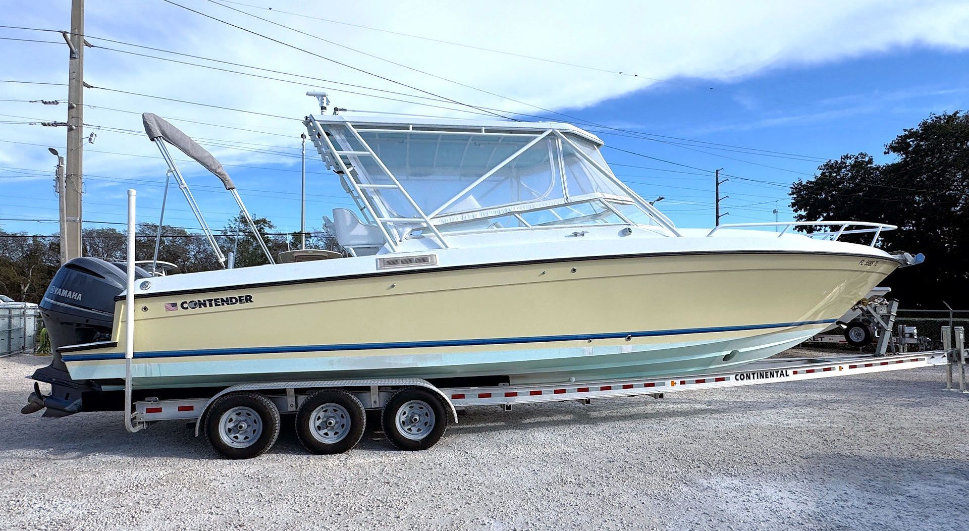 A light yellow and white center console boat on a triple-axle trailer, parked on a gravel lot under a blue sky.