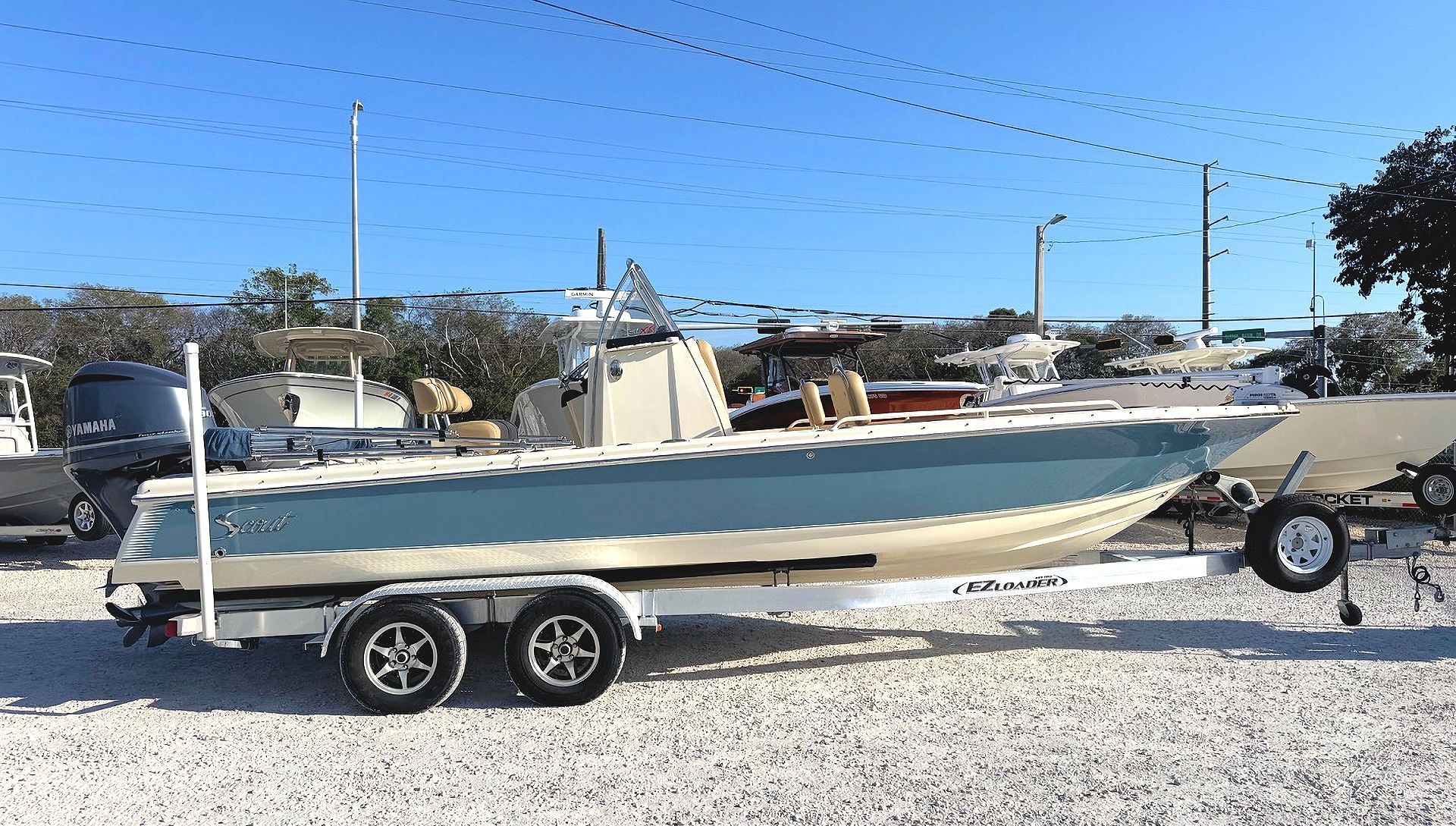 A teal and tan Scout bay boat on a tandem-axle trailer, parked on a gravel lot under a clear blue sky.