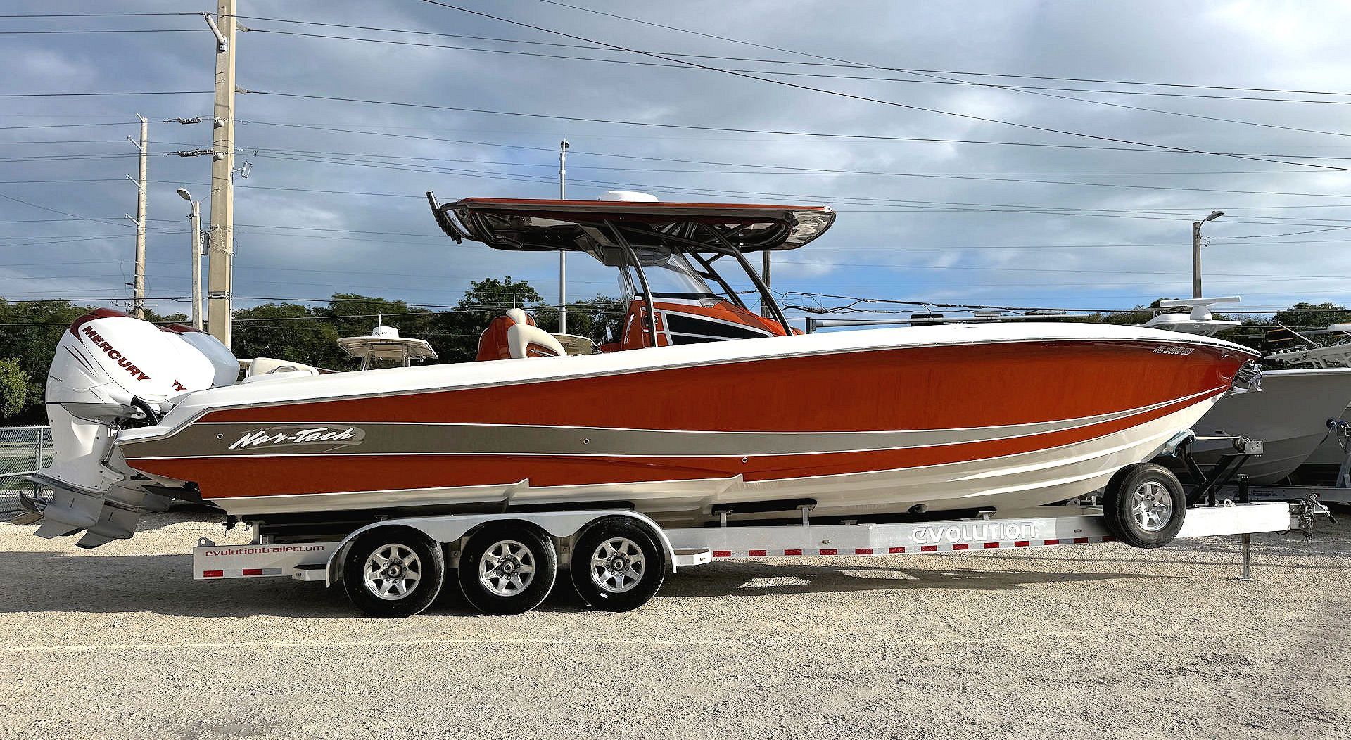 An orange and white center console boat on a triple-axle trailer, parked on a gravel lot under a cloudy sky.
