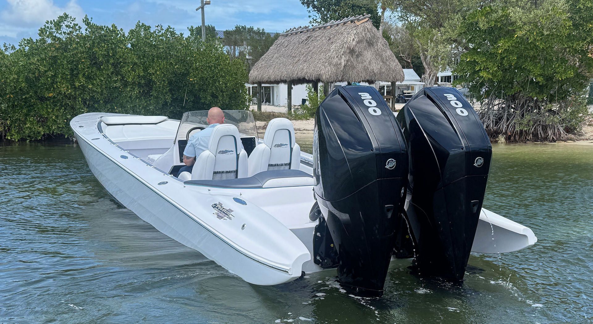 A white high-performance speedboat with dual black outboard motors anchored in shallow water near a grassy shoreline.