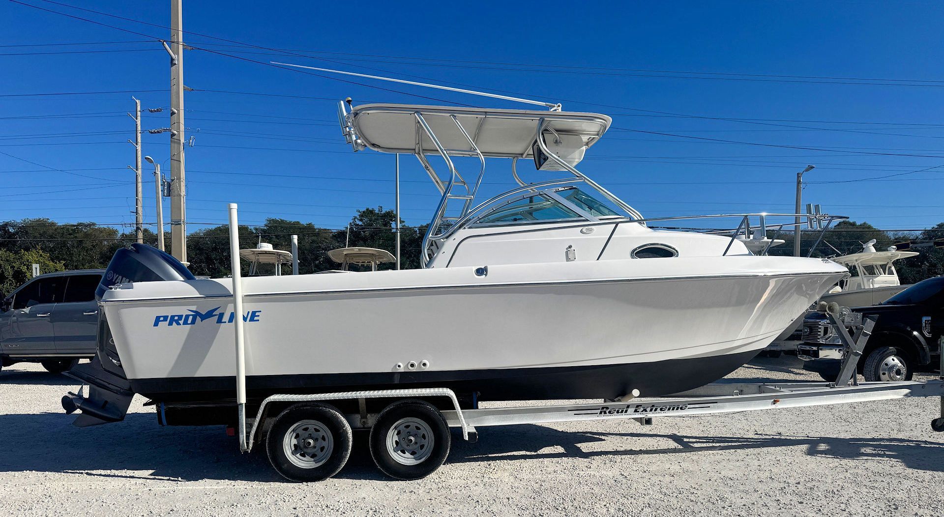 A white Pro-Line powerboat with a T-top on a dual-axle trailer, parked on a gravel lot under a clear blue sky.