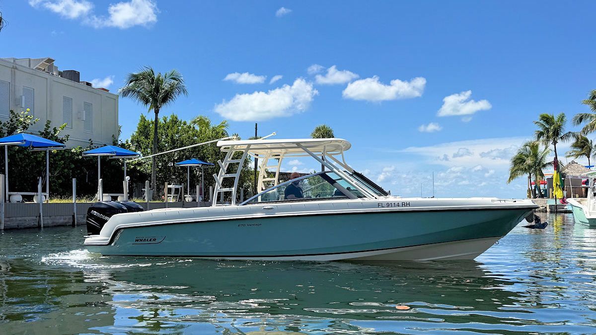 A light blue and white motorboat cruises slowly through a sunlit canal near palm trees and buildings under a blue sky.