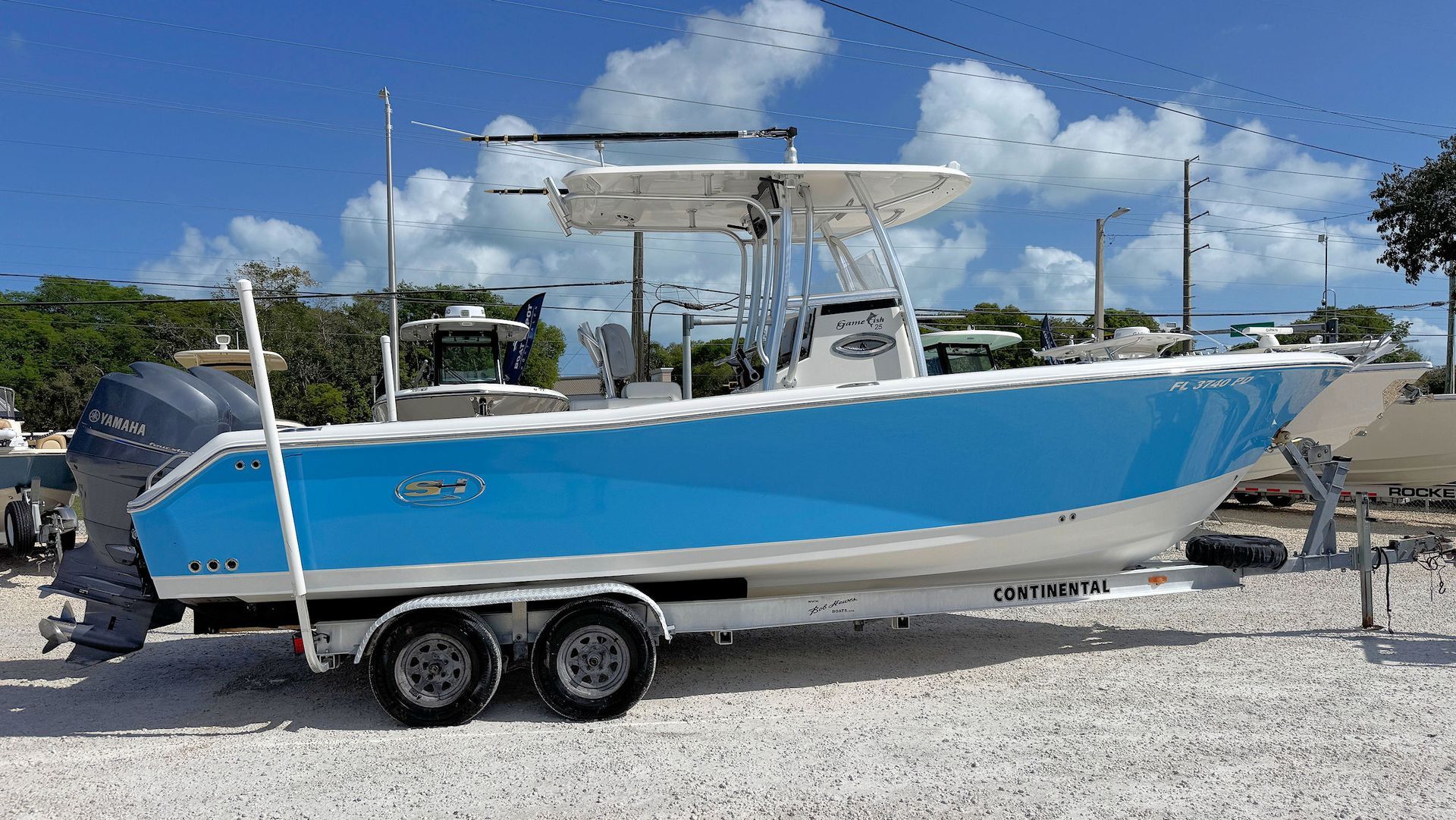 A light blue motorboat with a white center console sits on a double-axle trailer in a gravel lot under a sunny sky.