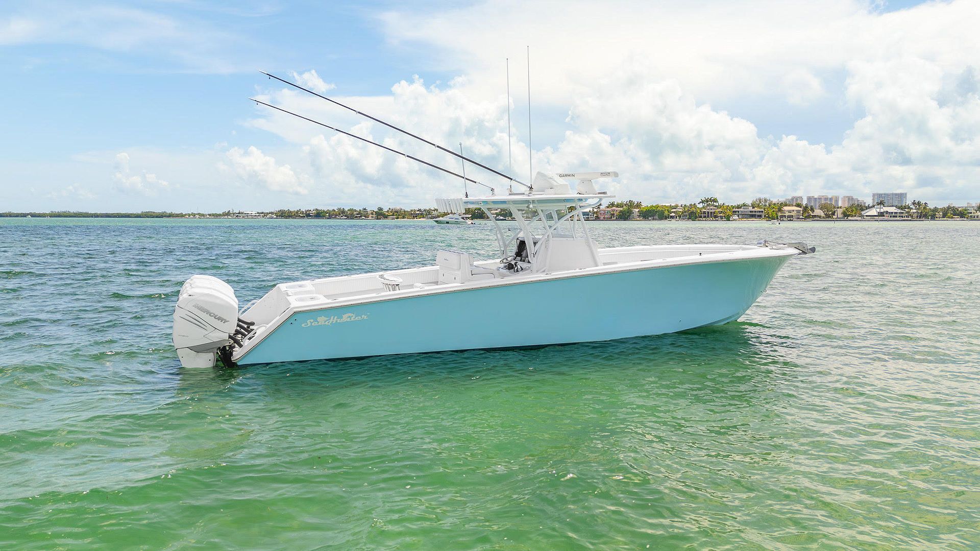 A light blue center console fishing boat floating on calm, clear water under a partly cloudy sky.