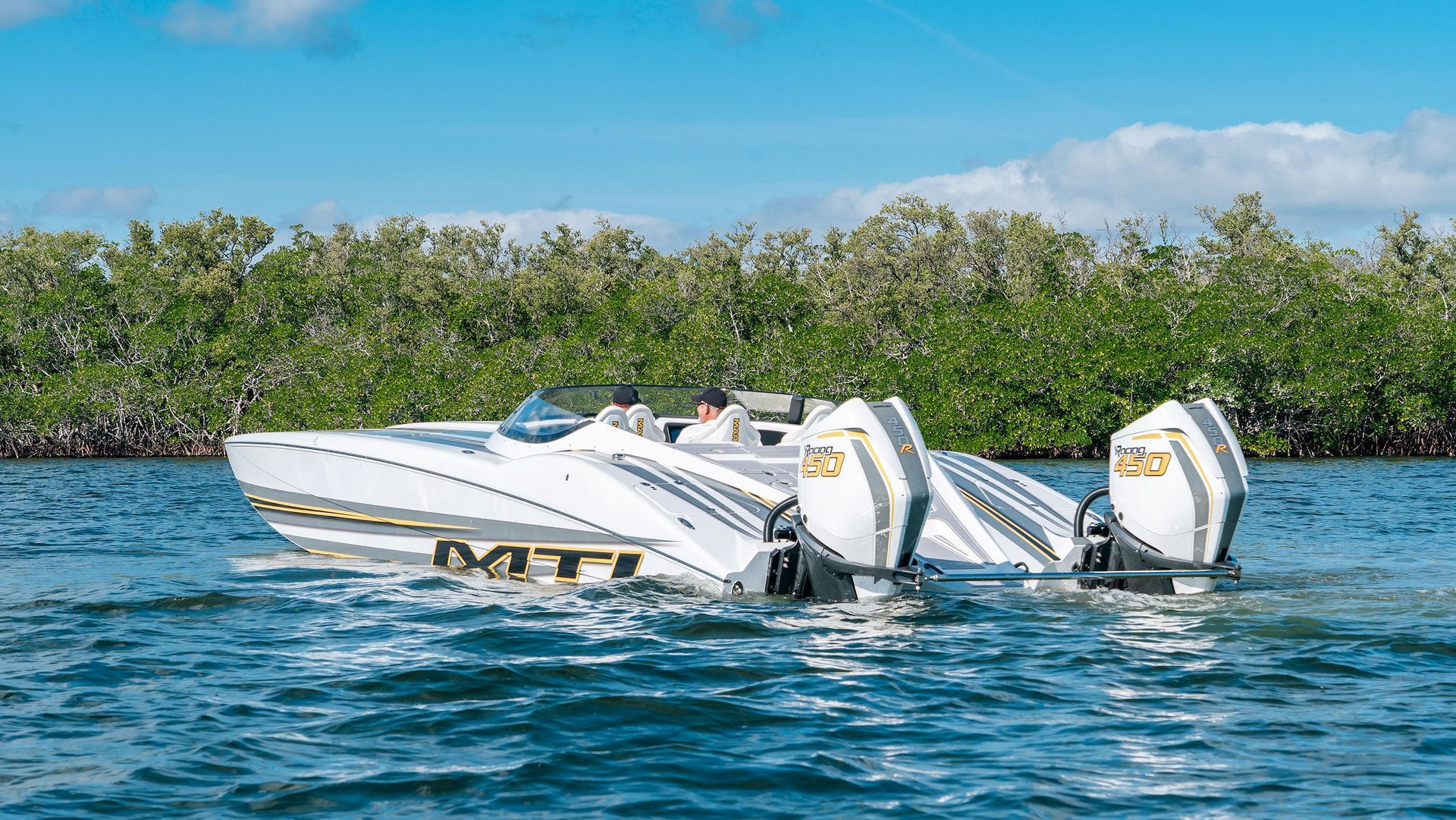 A white MTI high-performance catamaran powerboat with dual outboard motors cruising on blue water near a tree-lined shore.