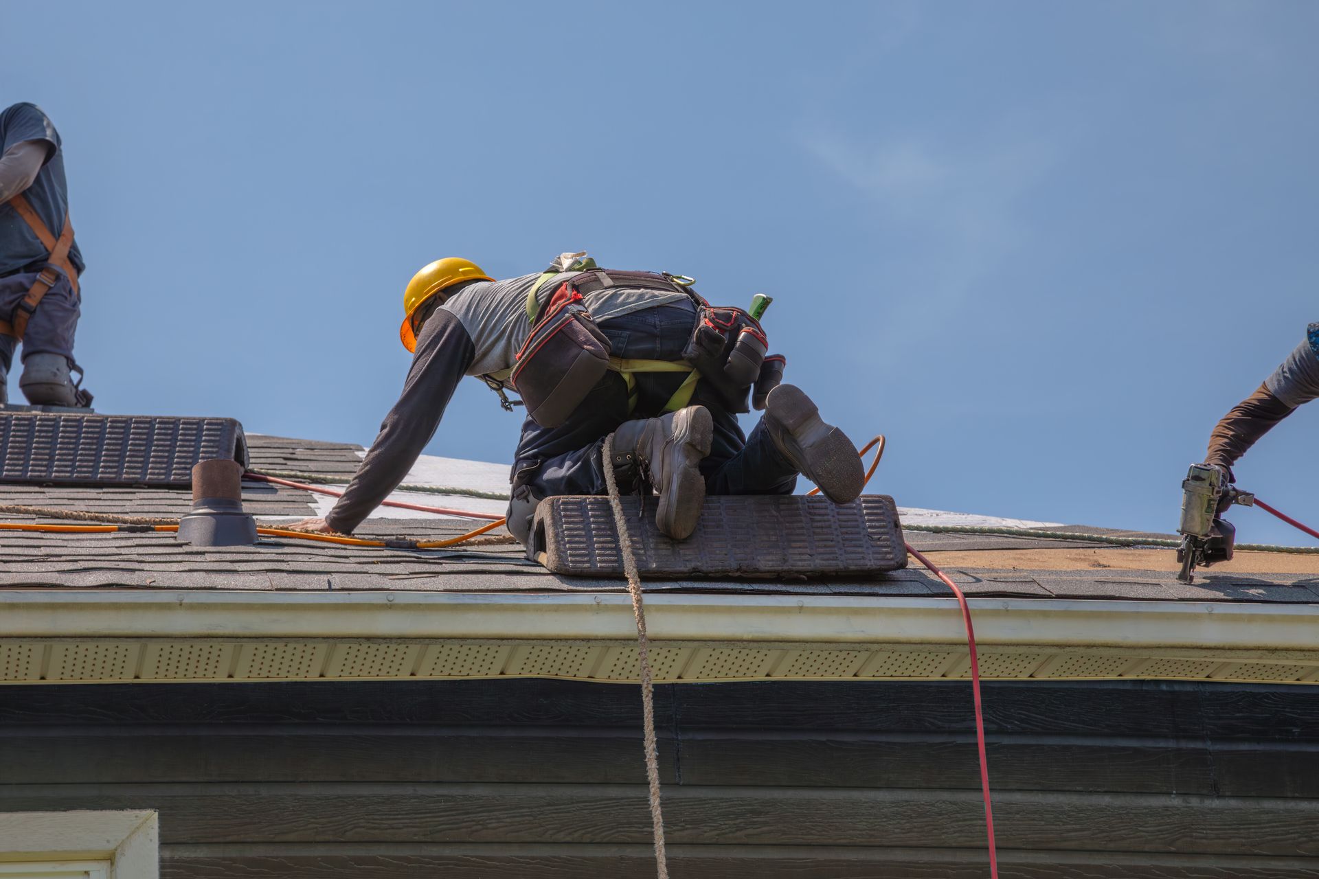 Roofers working on a rooftop under a blue sky, securing materials with ropes and tools.