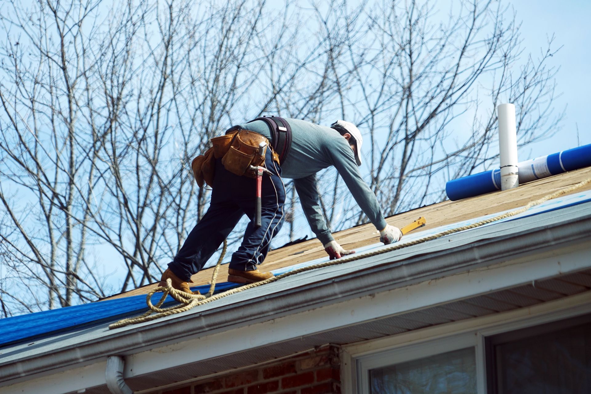 Worker on a roof securing shingles near a chimney, wearing a harness and tool belt.